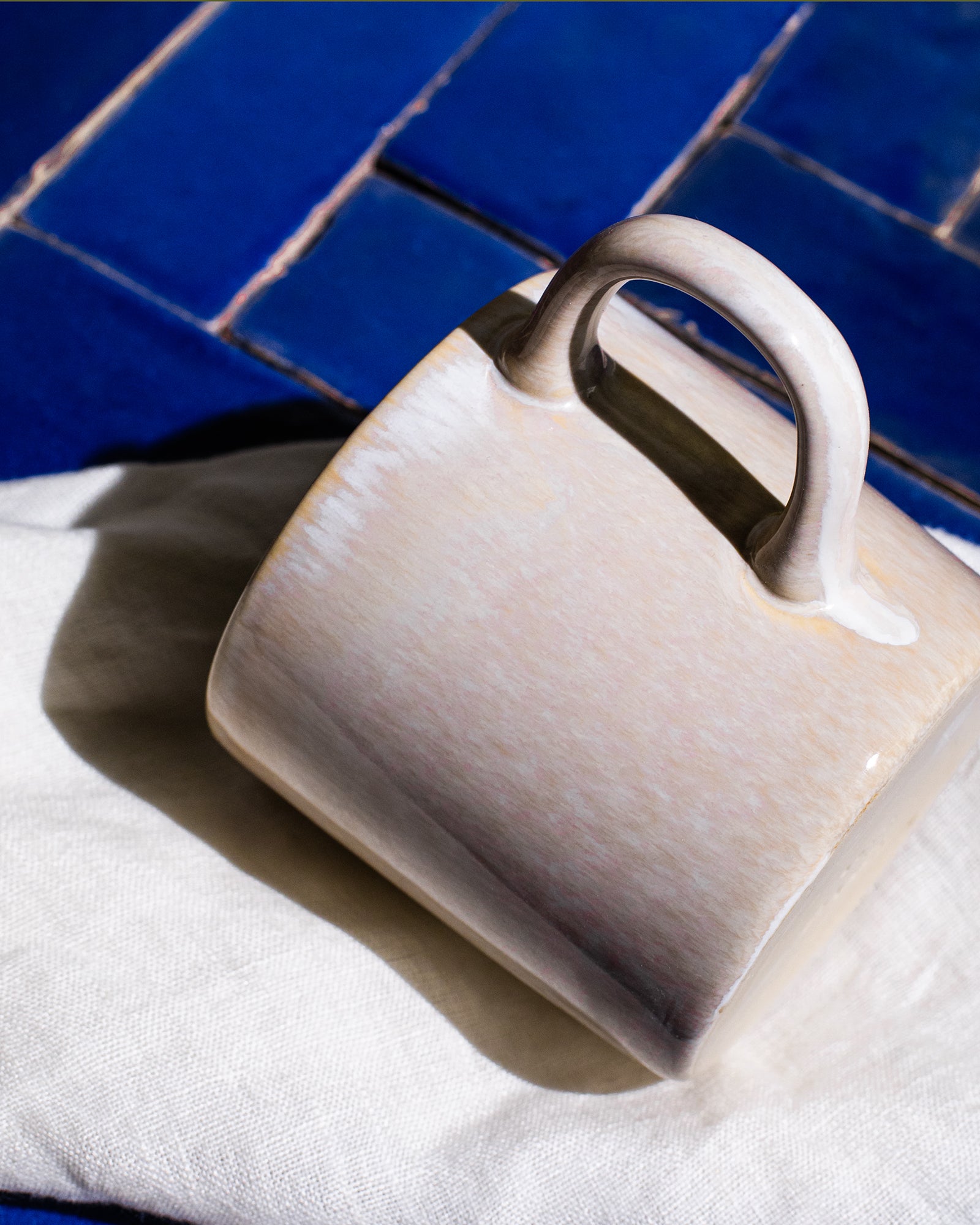 Keramiktasse liegt seitlich auf einem weißen Stofftuch vor einem Hintergrund aus blauen Fliesen.