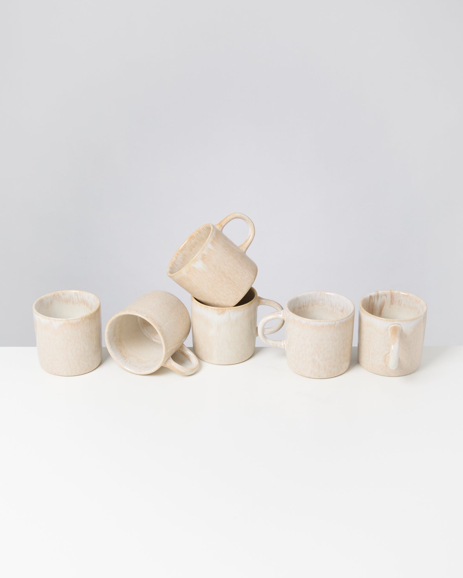 Sechs cremefarbene Tassen stapeln sich unordentlich auf einem weißen Tisch vor einem grauen Hintergrund.