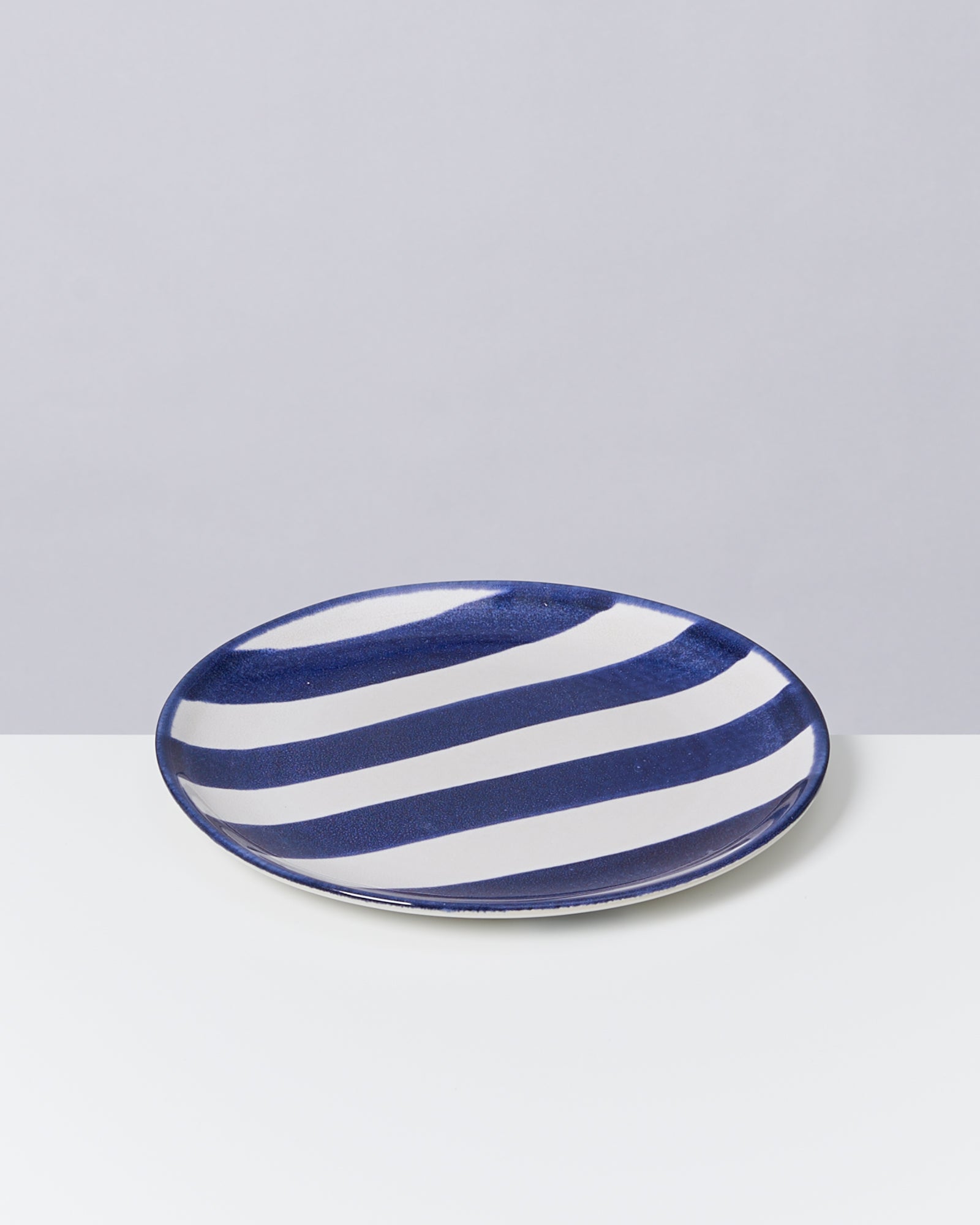 Blauer Teller mit weißen, diagonalen Streifen auf einer weißen Oberfläche, vor grauem Hintergrund.