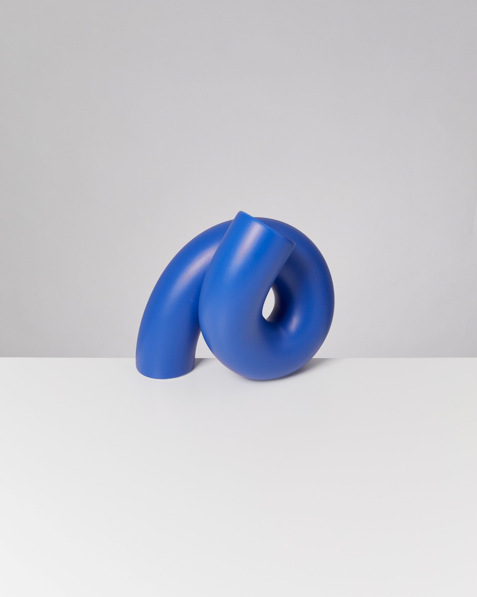 Blaue, abstrakte Skulptur mit geschwungenen Formen auf weißer Oberfläche vor grauem Hintergrund.