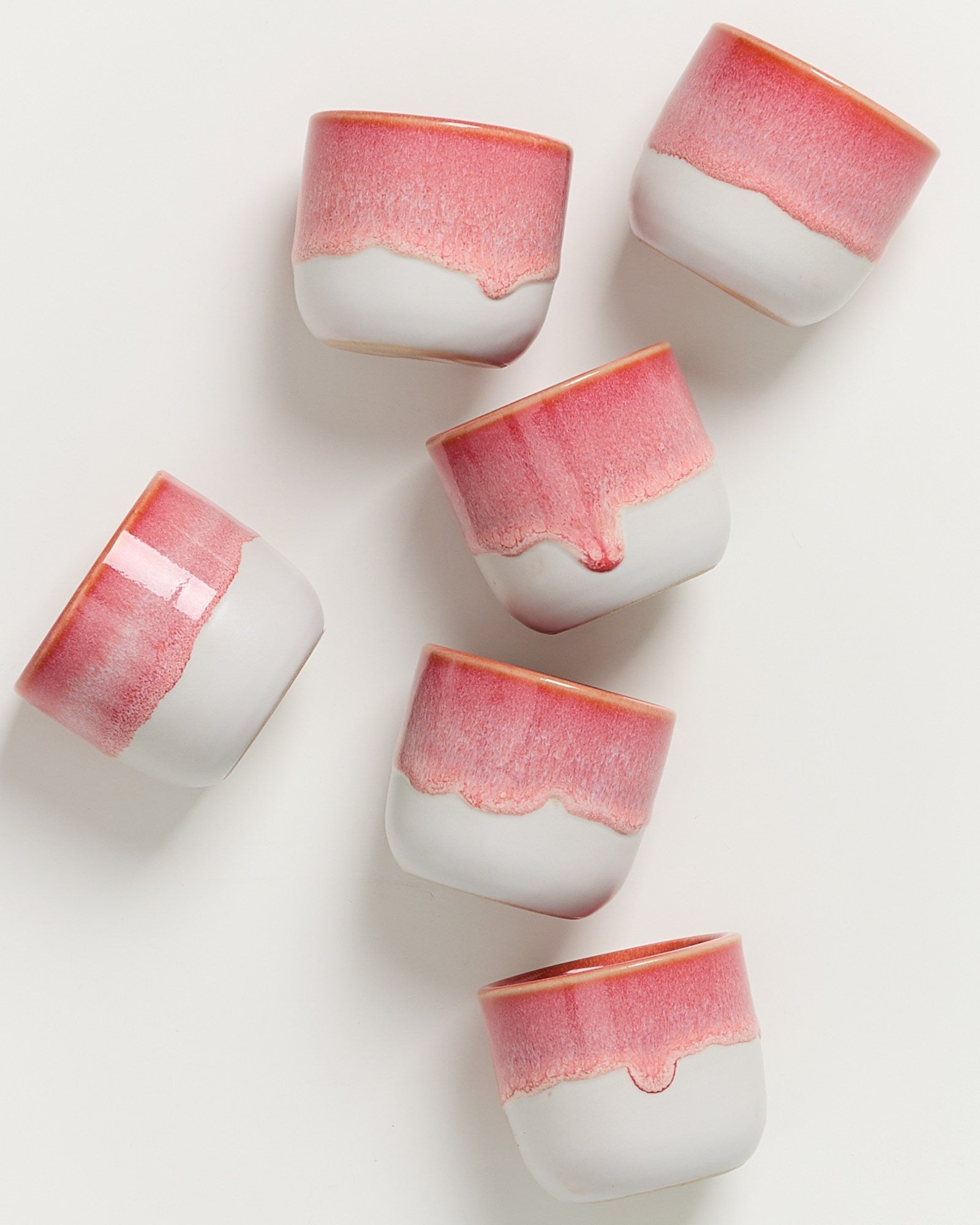 Sechs weiße Tassen mit rosa Rand und unregelmäßigem Muster liegen auf einem weißen Hintergrund verstreut.