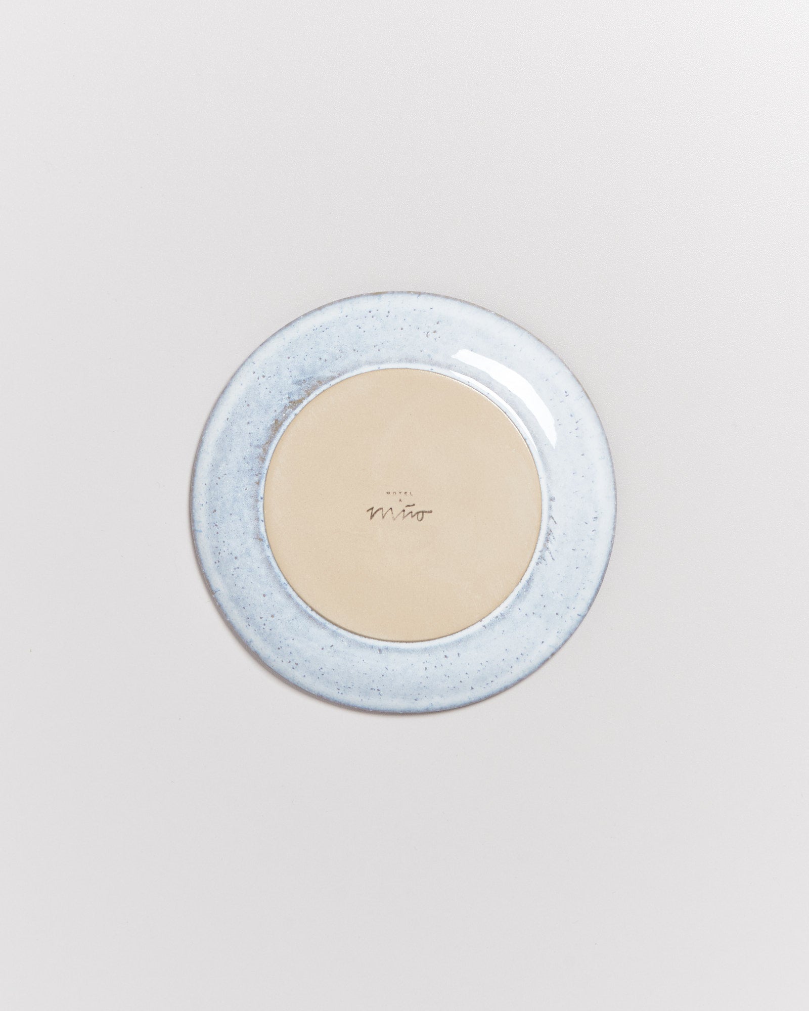 Hellblaue, runde Keramikplatte mit einem beigen Zentrum und einem kleinen Logo in der Mitte, auf hellem Hintergrund.