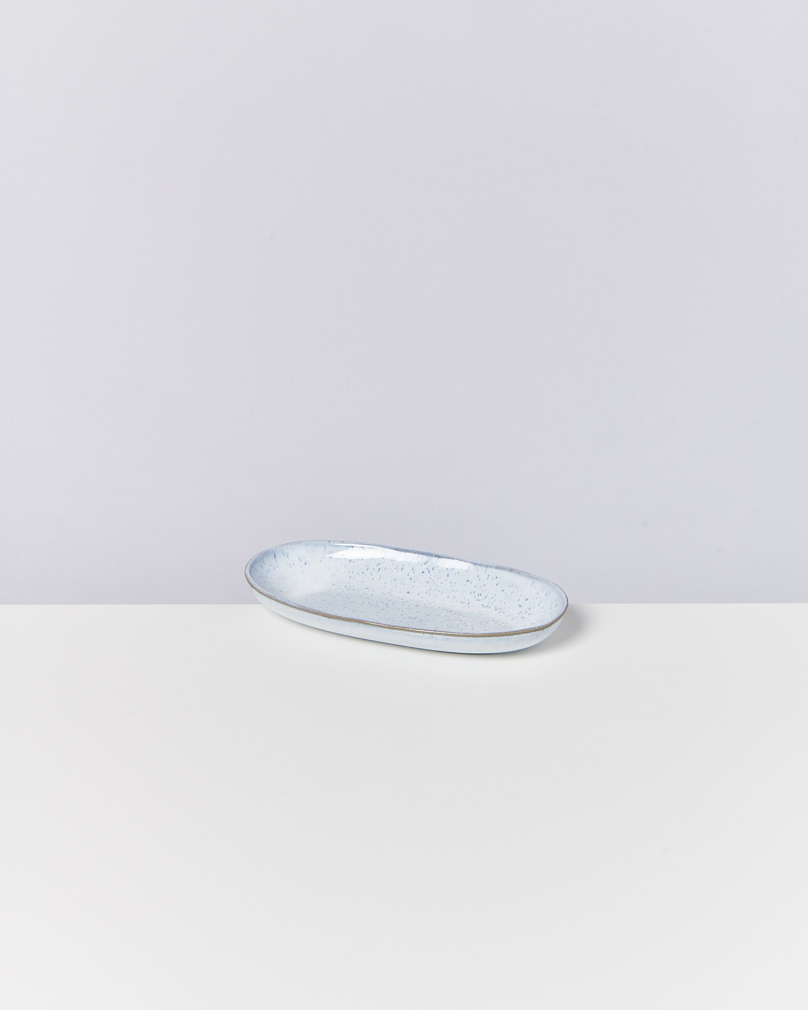Ein ovaler, blaugrau gesprenkelter Keramikteller auf einem weißen Tisch vor einem grauen Hintergrund.