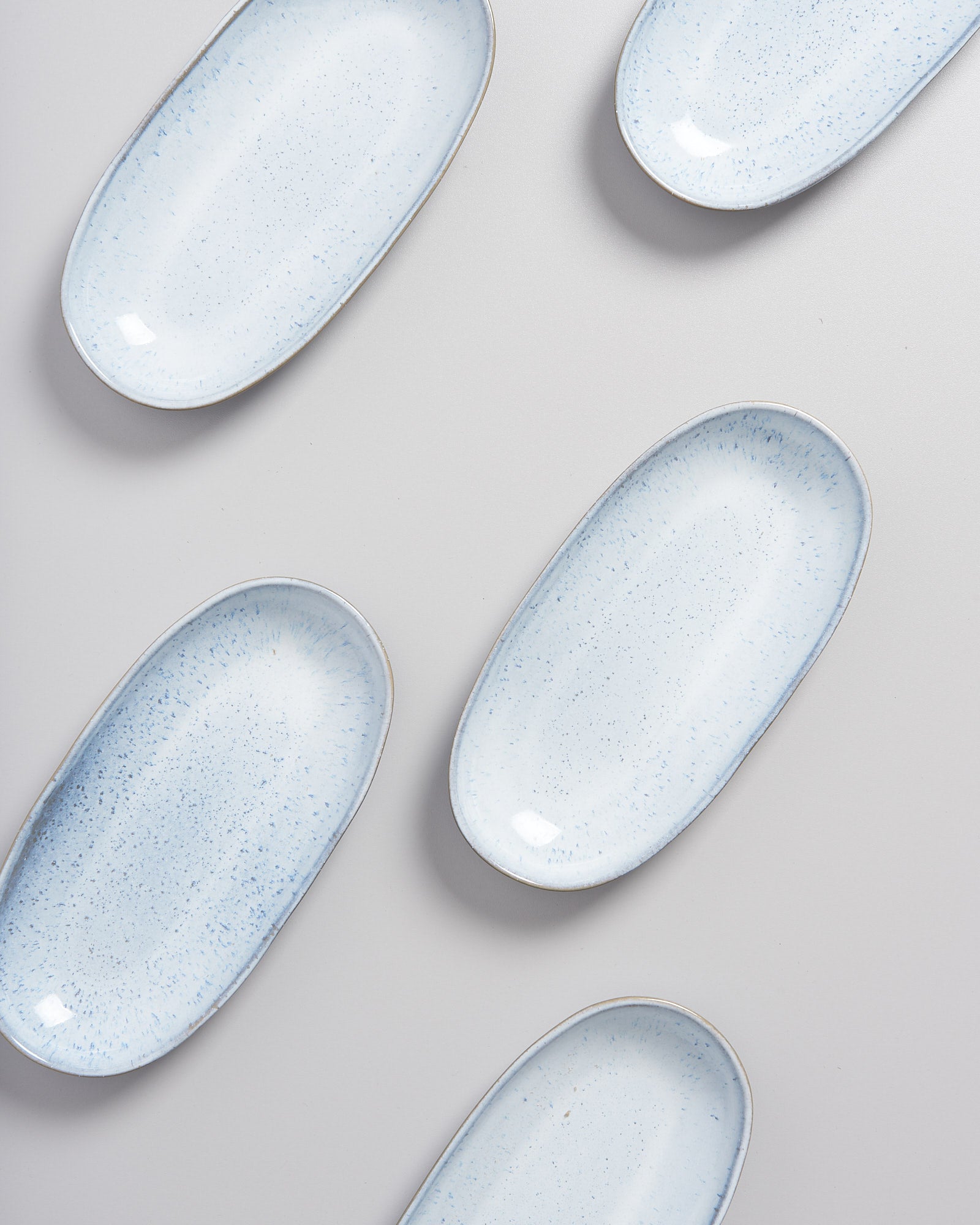 Sechs ovale, hellblaue Keramikteller mit Sprenkeln sind auf einer hellgrauen Oberfläche verteilt.