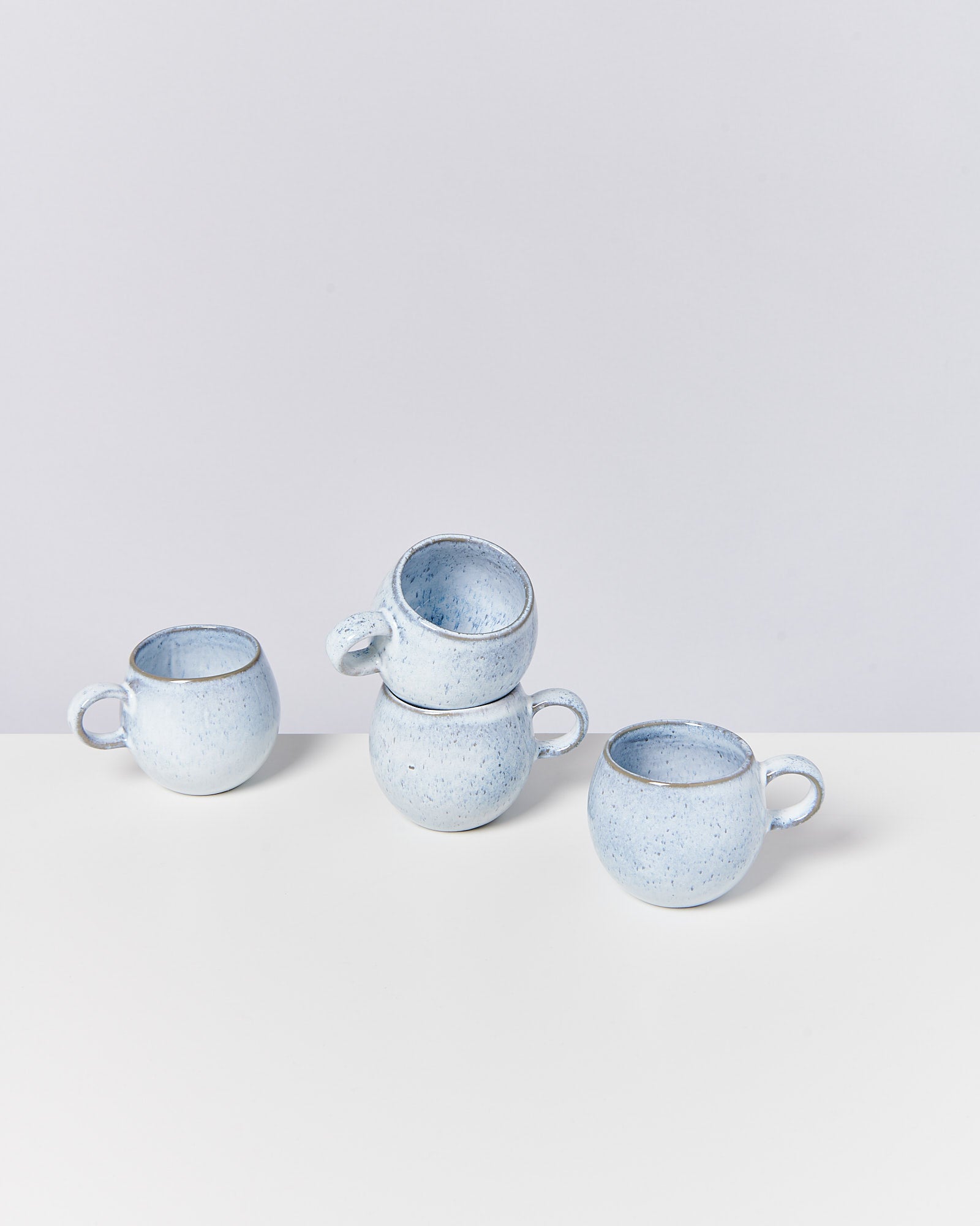 Vier blaue Keramiktassen mit Henkel, eine Tasse ist schief auf zwei anderen gestapelt, auf einem weißen Tisch.