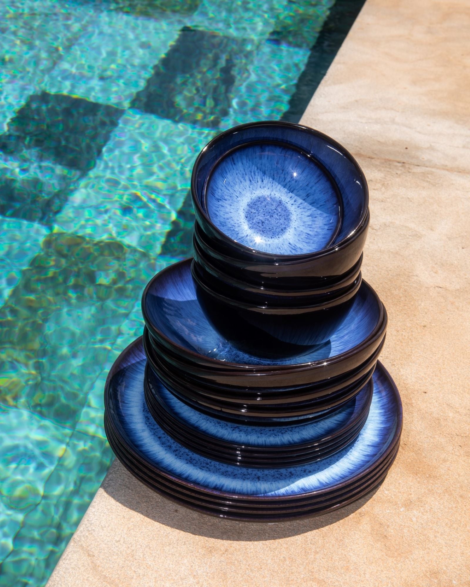 Ein Stapel blauer Keramikteller steht auf hellem Boden neben einem Pool mit blau-grün schimmerndem Wasser.