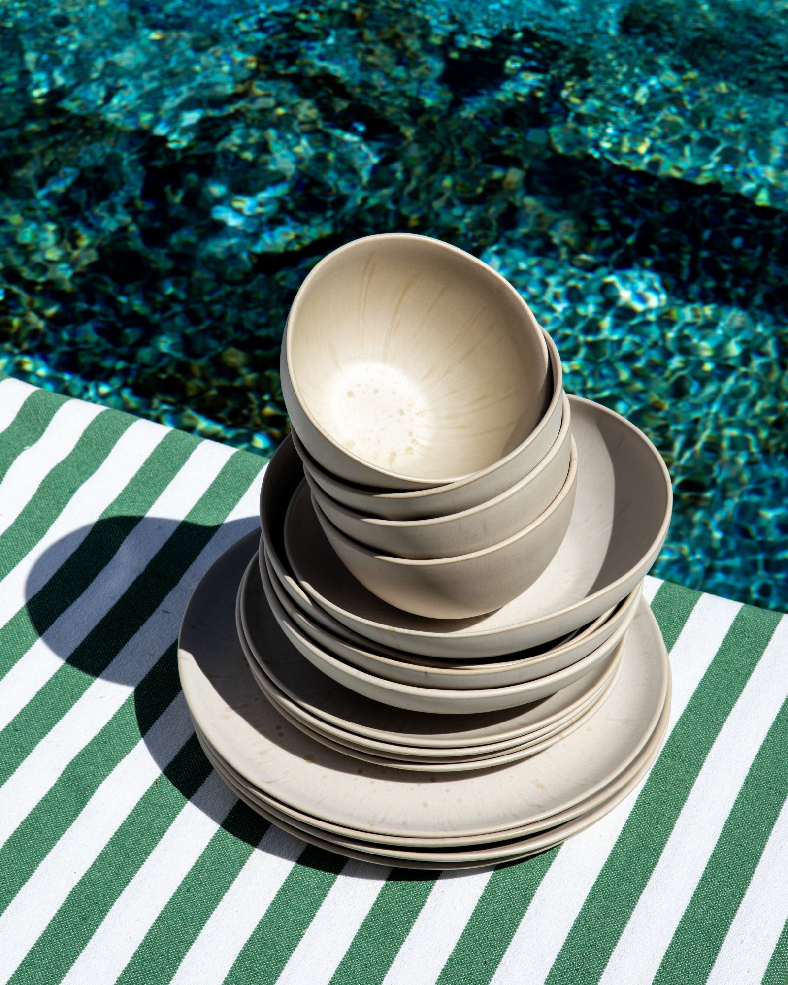 Ein Stapel heller Schalen und Teller steht auf einem gestreiften grünen und weißen Tuch neben einem Pool.