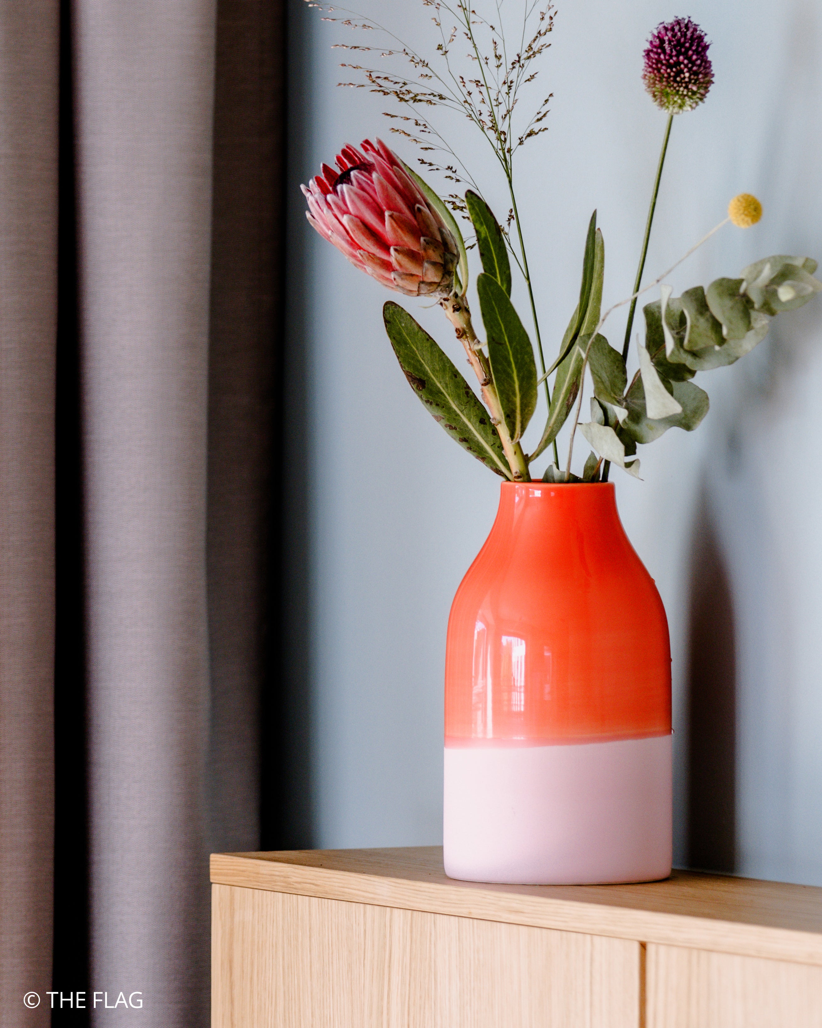 Ein orange-pinker Keramikvase mit exotischen Blumen steht auf einem Holztisch vor einem blauen Hintergrund und Vorhang.