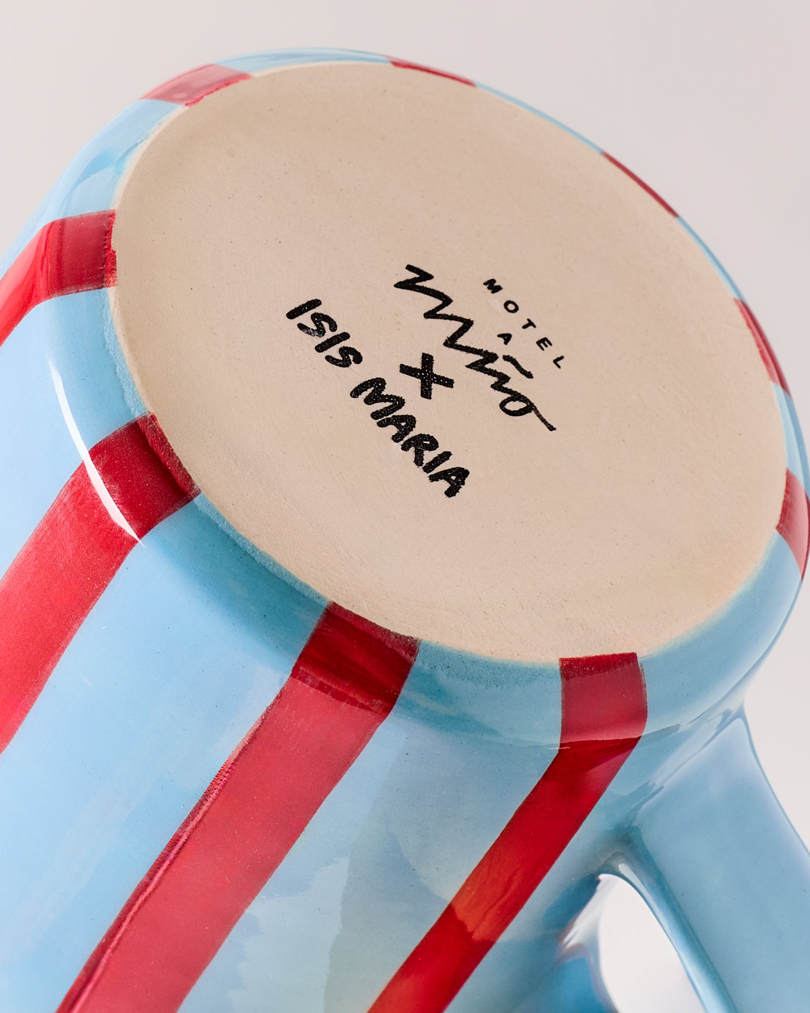 Nahaufnahme eines blauen Bechers mit roten Streifen und der Aufschrift "Motel a Miio x Isis Maria" auf der Unterseite.