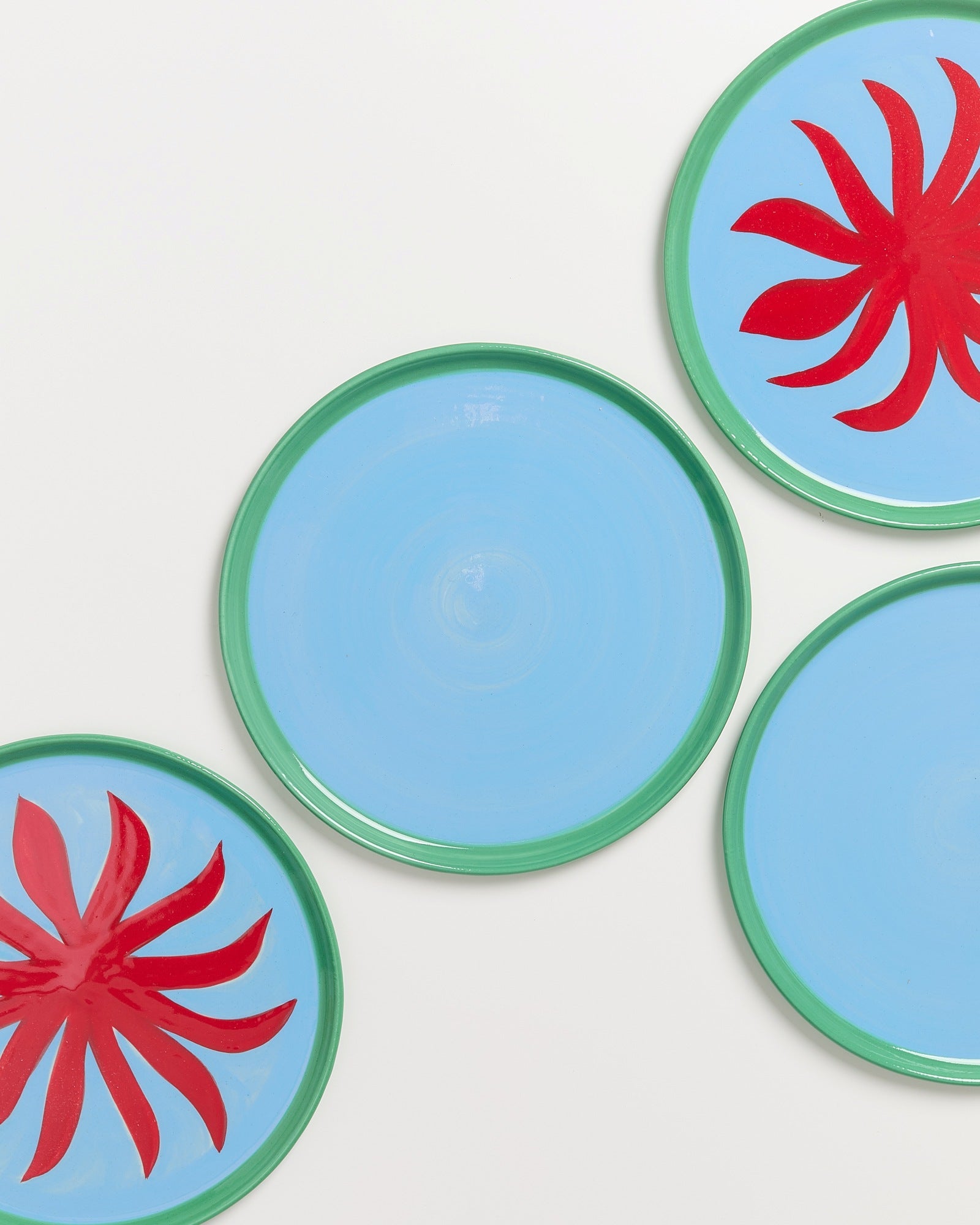 Vier blaue Teller mit grünem Rand, zwei mit roten Blumendesigns, auf weißem Hintergrund.