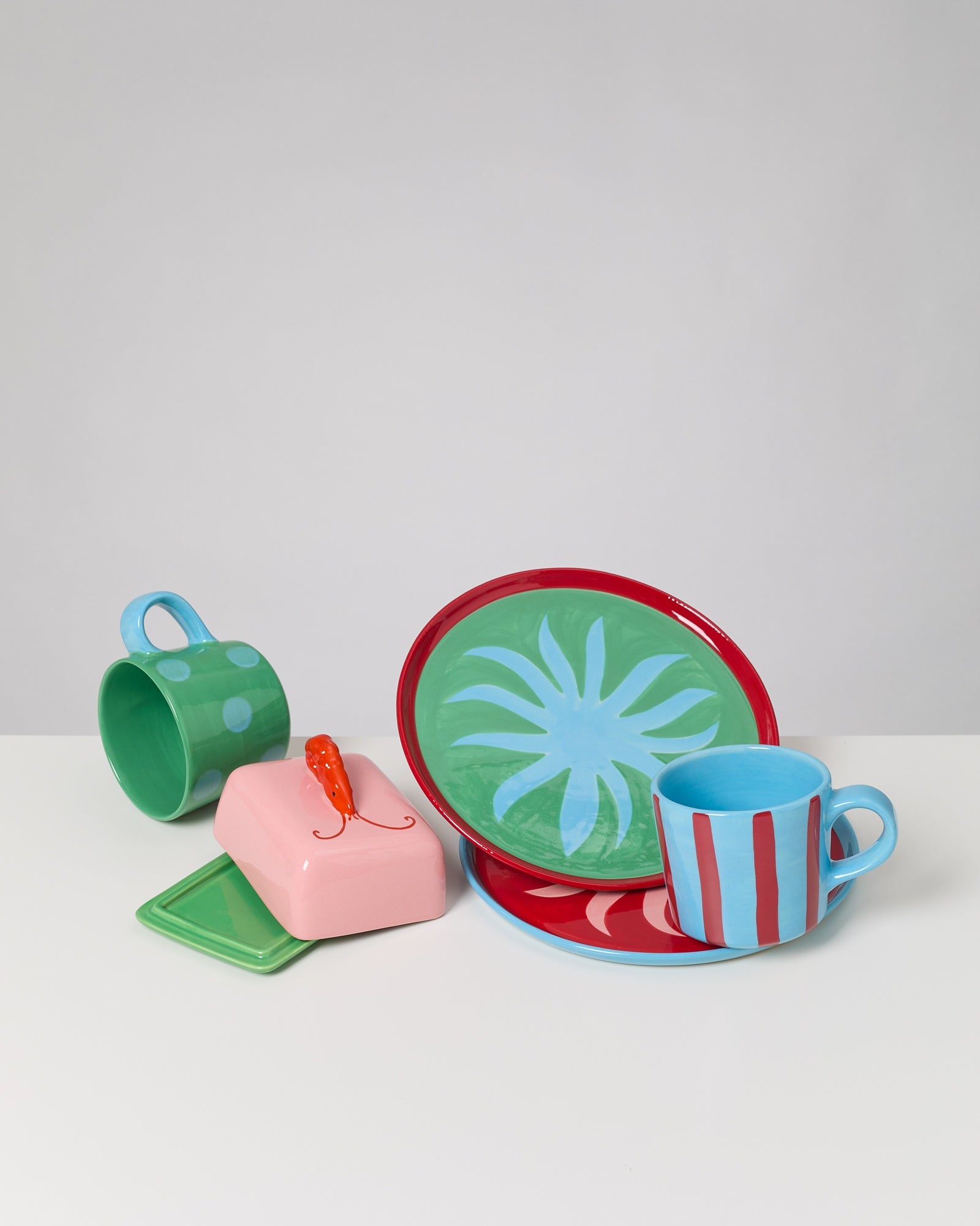 Buntes Geschirr-Set mit zwei Tassen, zwei Tellern und einer rosa Butterdose mit einem orangefarbenen Griff auf einem Tisch.