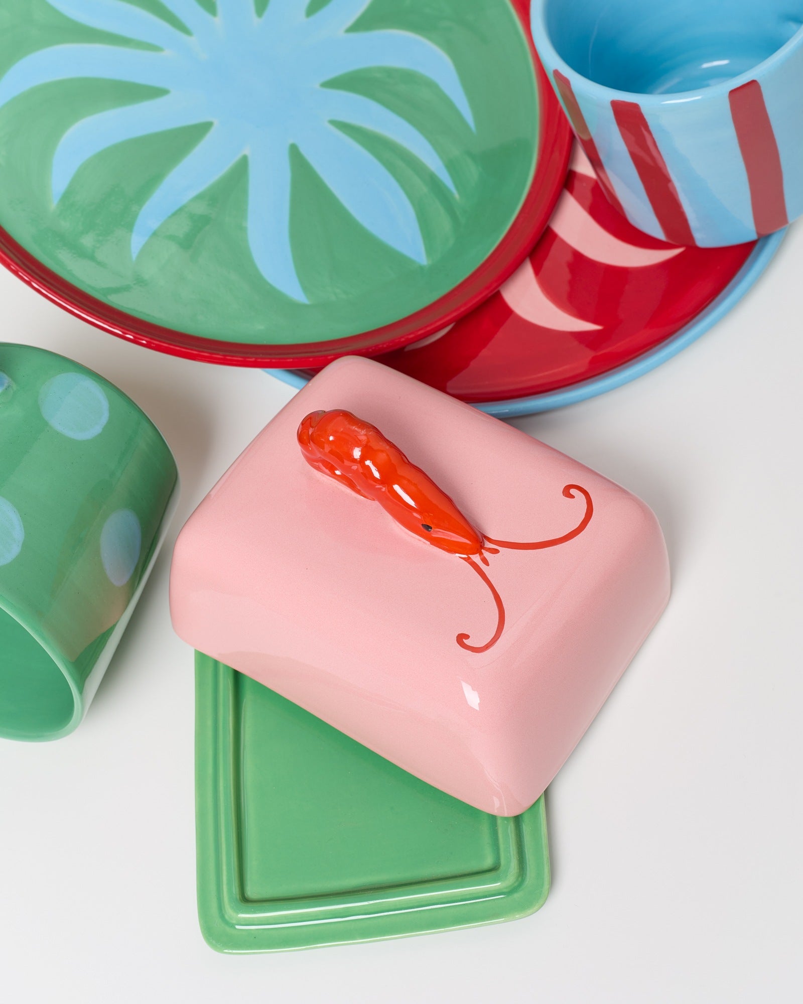 Bunte Keramikgeschirr mit einem rosa Behälterdeckel, verziert mit einer orangen Garnele, auf grünen und roten Tellern.
