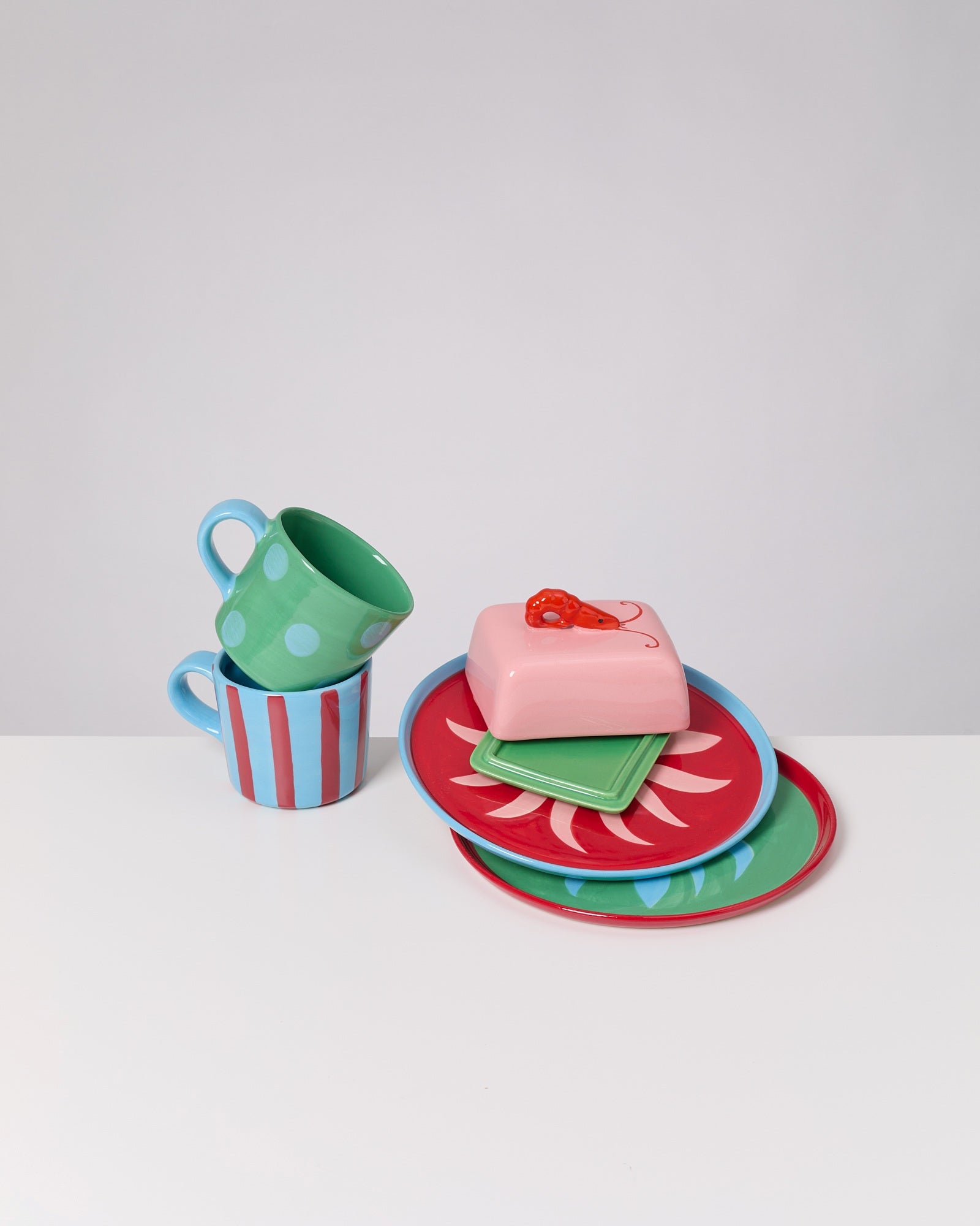 Zwei bunte Tassen neben einem Stapel farbenfroher Teller und einer rosa Butterdose mit Kirschenmuster.