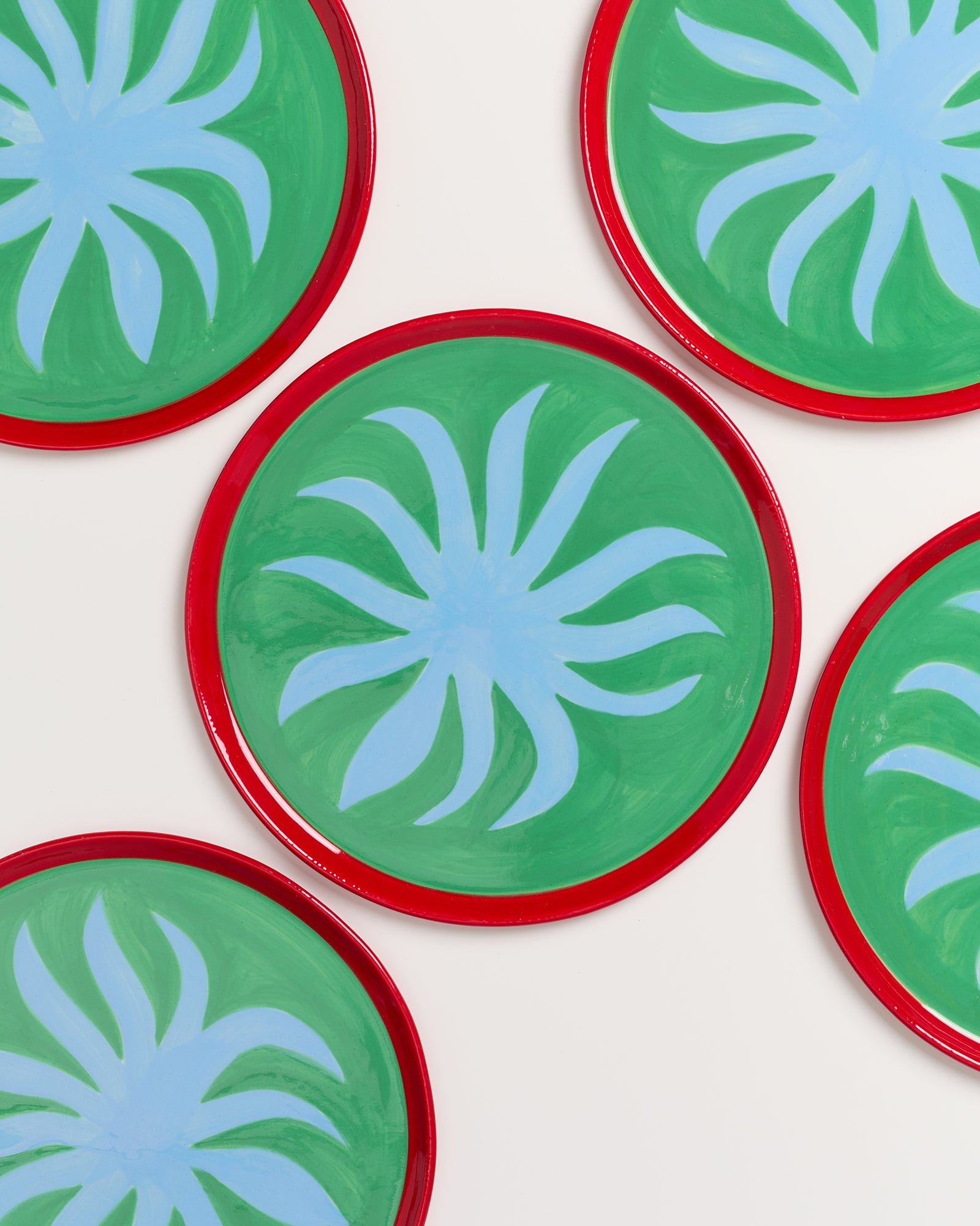 Fünf runde Teller mit grünem Hintergrund, blauen Mustern und roten Rändern liegen auf einem weißen Untergrund.