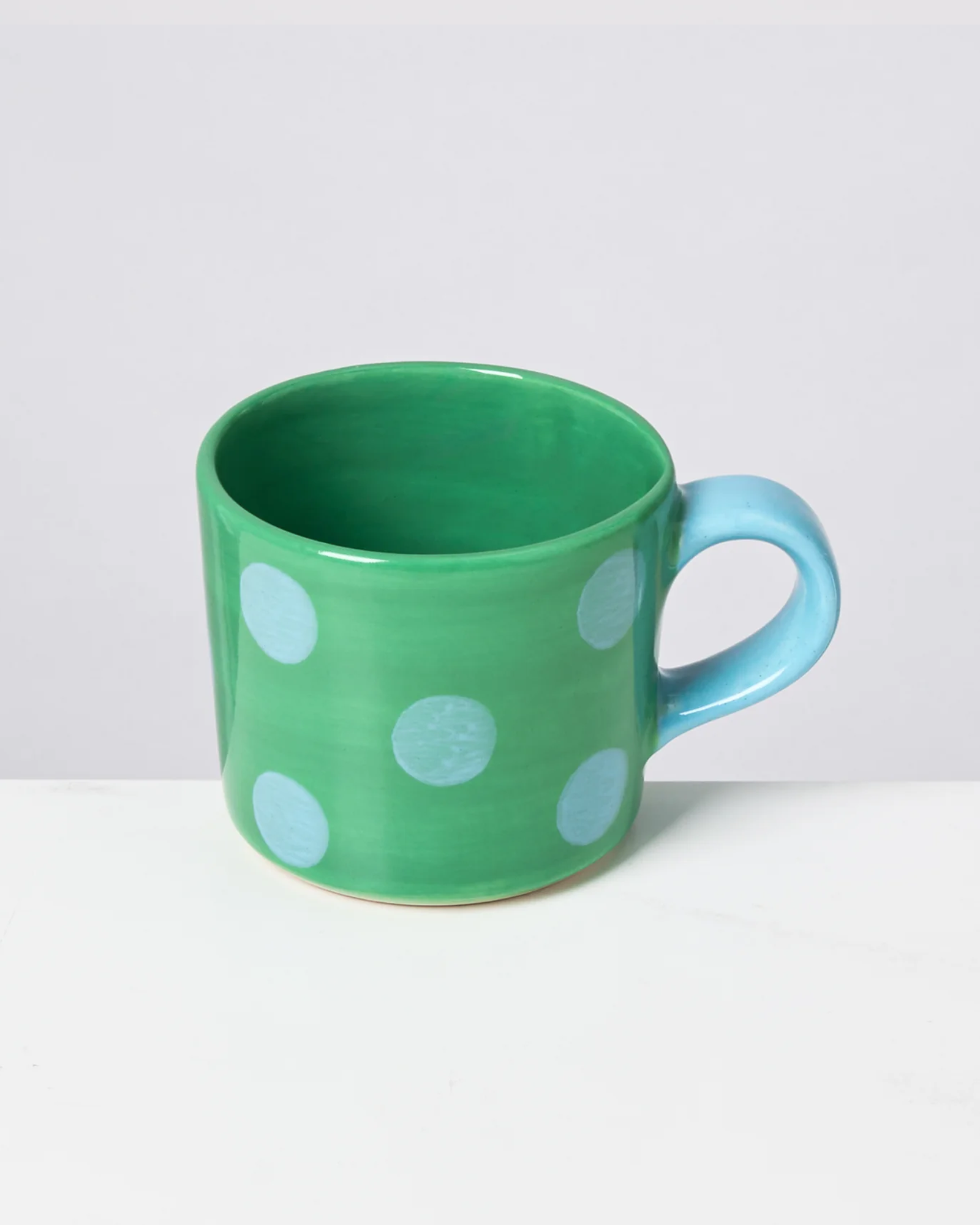 Grüne Tasse mit blauen Punkten und blauem Henkel vor einem weißen Hintergrund.