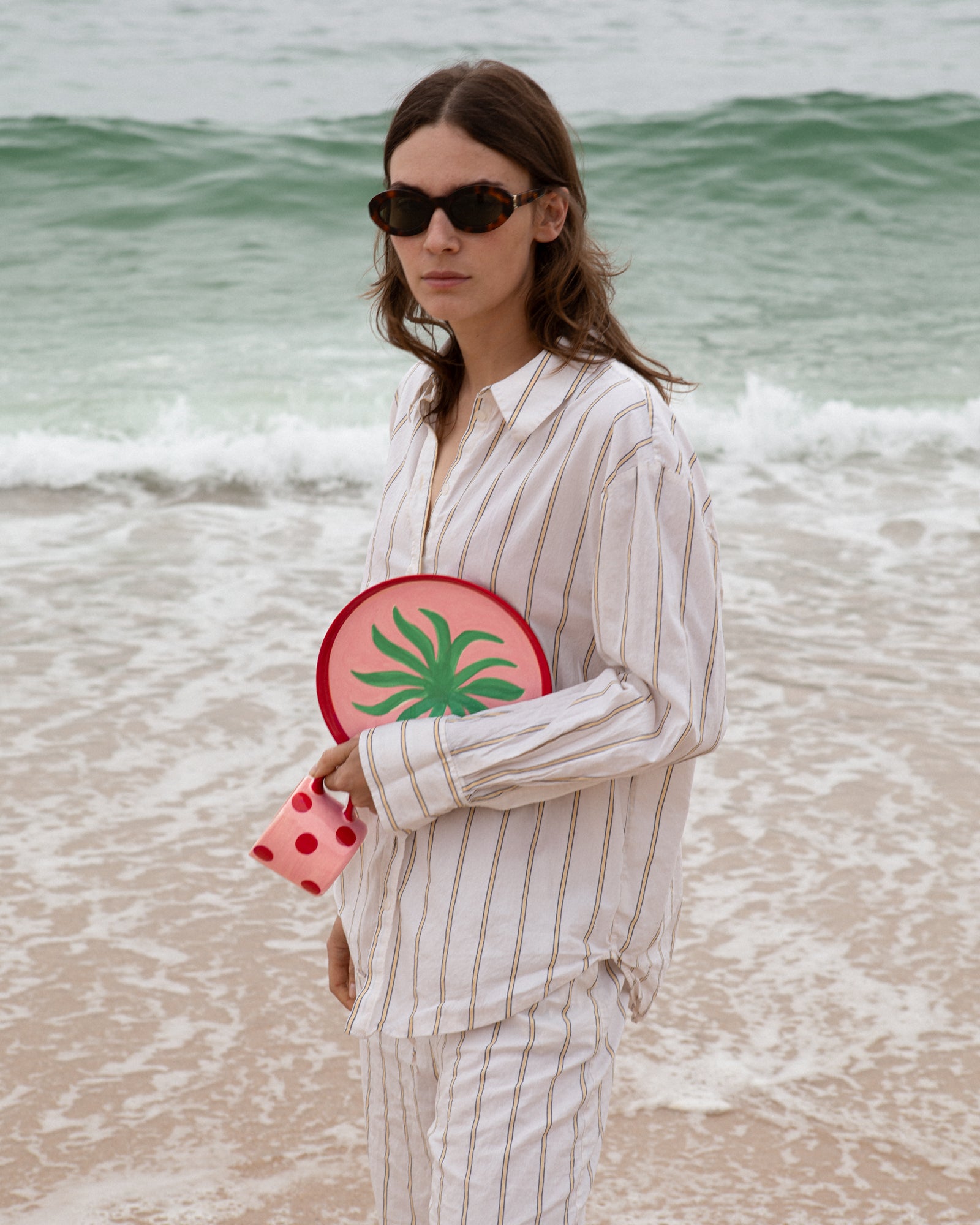 Eine Frau in gestreifter Kleidung steht am Strand mit einer rot-grünen Strandtasche und trägt Sonnenbrillen.