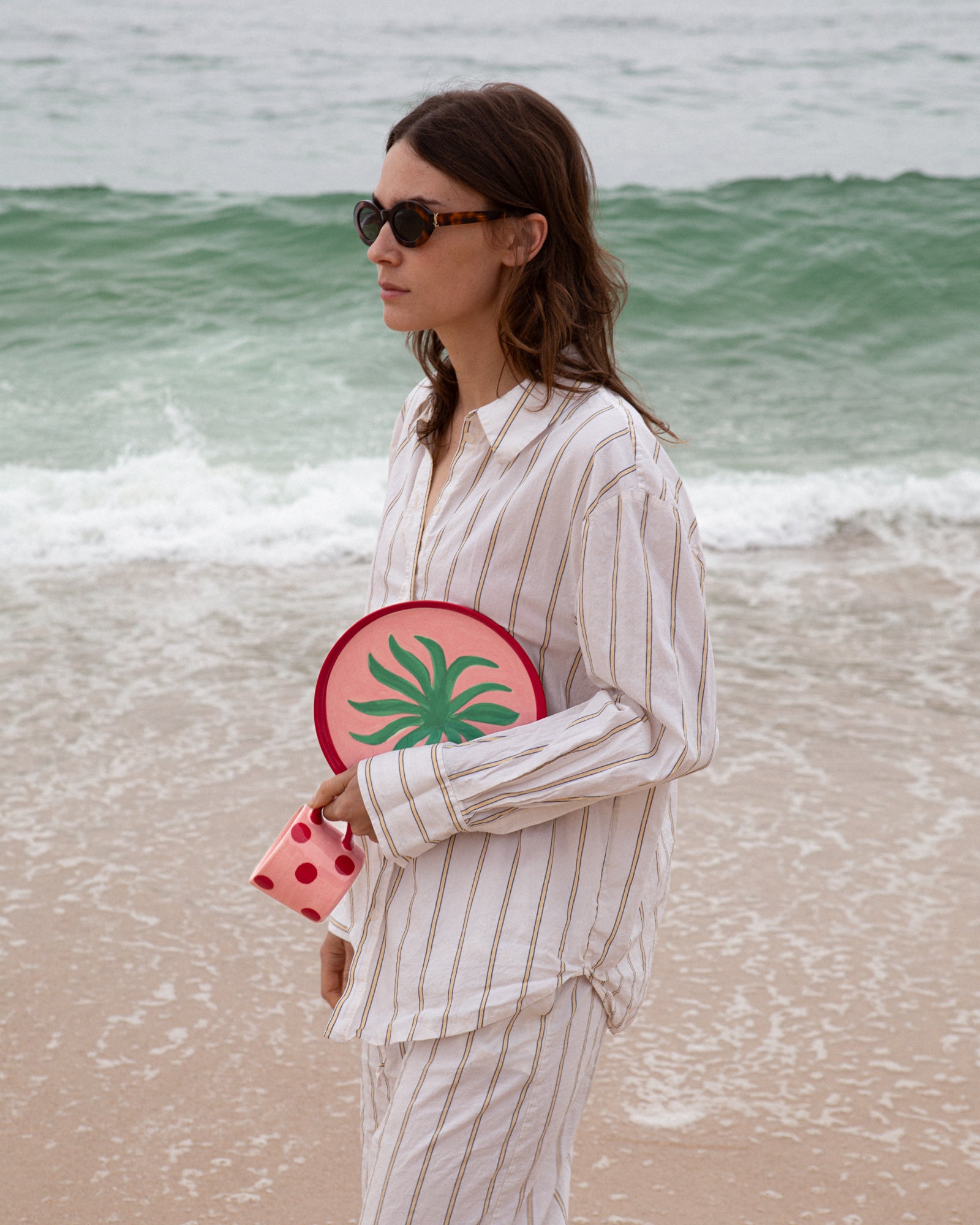 Frau in gestreiftem Hemd und Sonnenbrille steht am Strand, hält einen runden Gegenstand mit grünem Muster und roten Tassen.