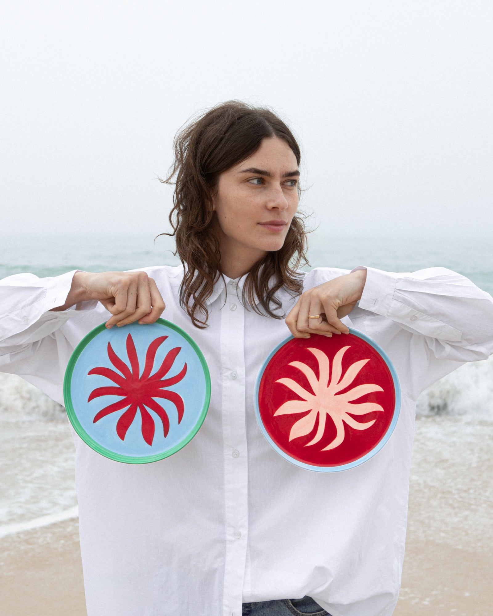 Eine Frau am Strand hält zwei bunte Teller mit spiralförmigen Mustern in die Kamera.