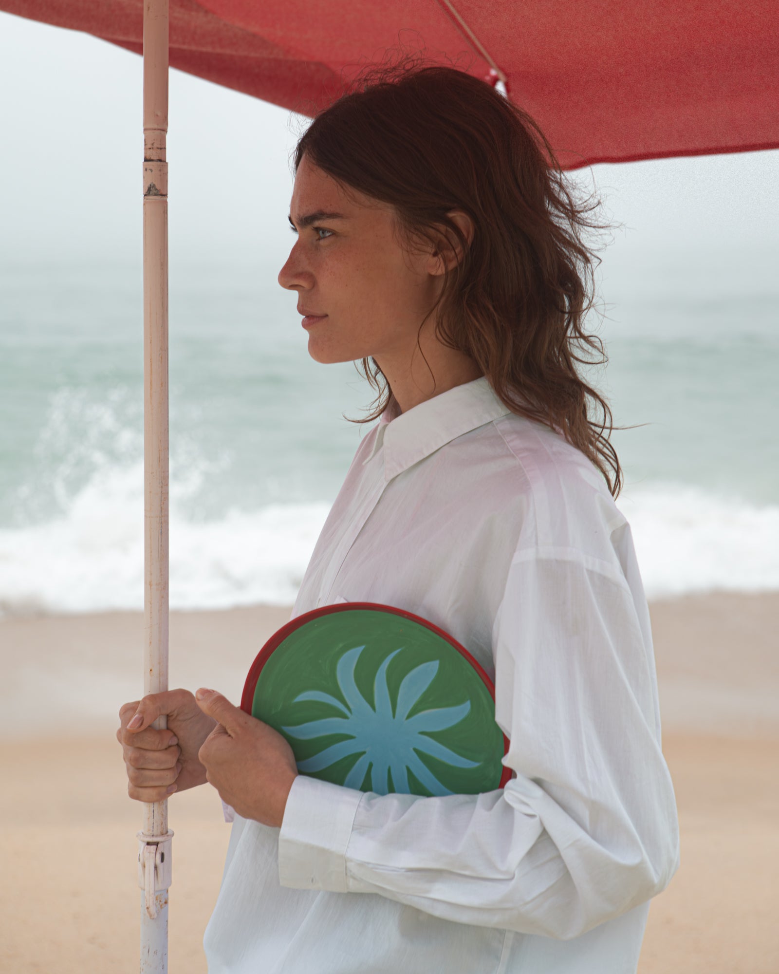 Frau unter rotem Sonnenschirm am Strand, in weißem Hemd, hält ein grünes rundes Objekt mit blauem Muster.