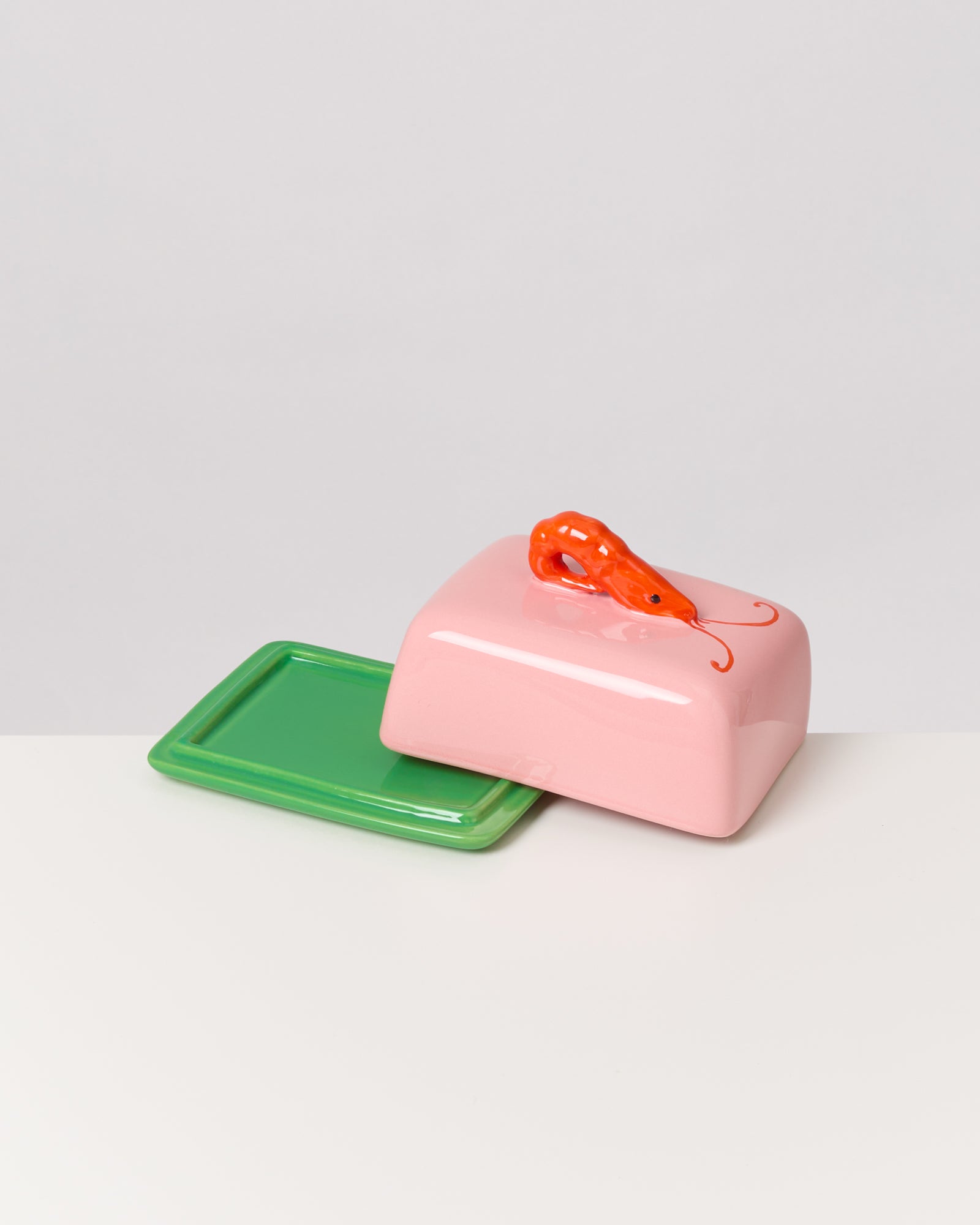 Rosa Butterdose mit einem orangefarbenen Garnelen-Dekor auf einem grünen Tablett auf weißem Hintergrund.