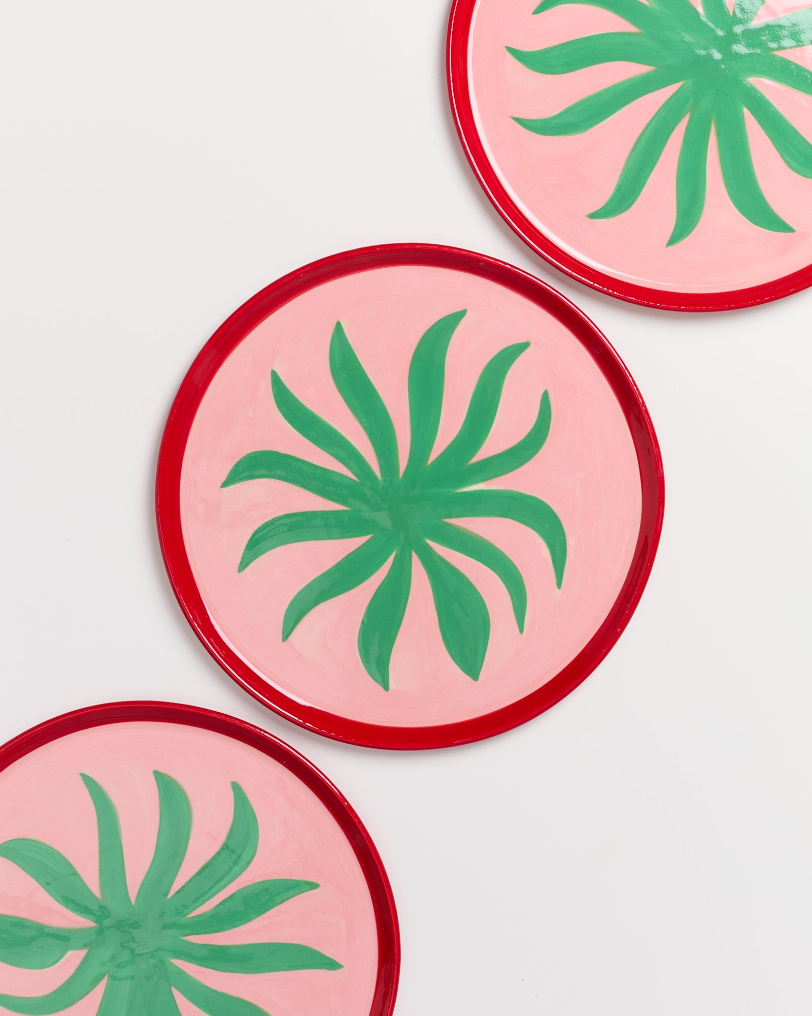 Drei runde Teller mit grünem Blattmuster auf rosa Hintergrund und rotem Rand.
