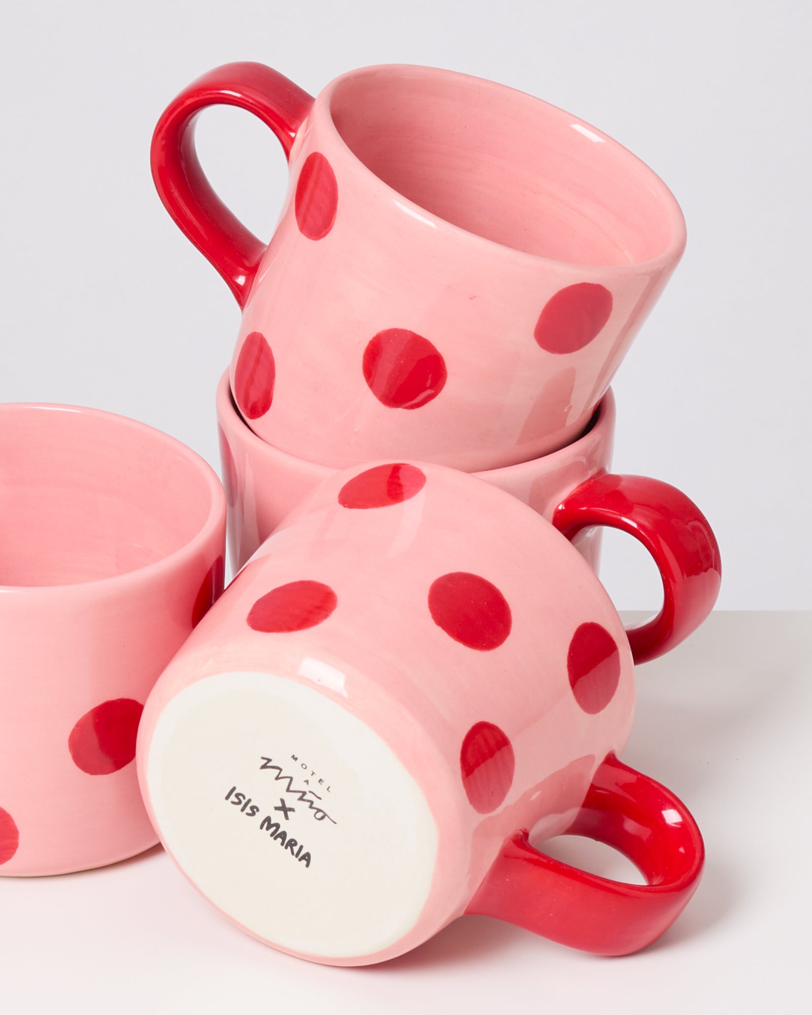 Vier rosa Tassen mit roten Punkten und Griffen sind gestapelt, mit einer Markierung "ISIS MARIA" auf der Unterseite.