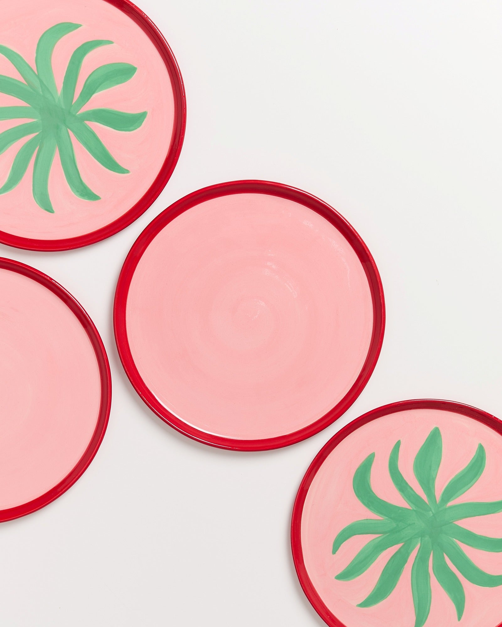 Vier rosa Teller mit rotem Rand, zwei davon mit grünen, palmenartigen Mustern auf hellem Hintergrund.