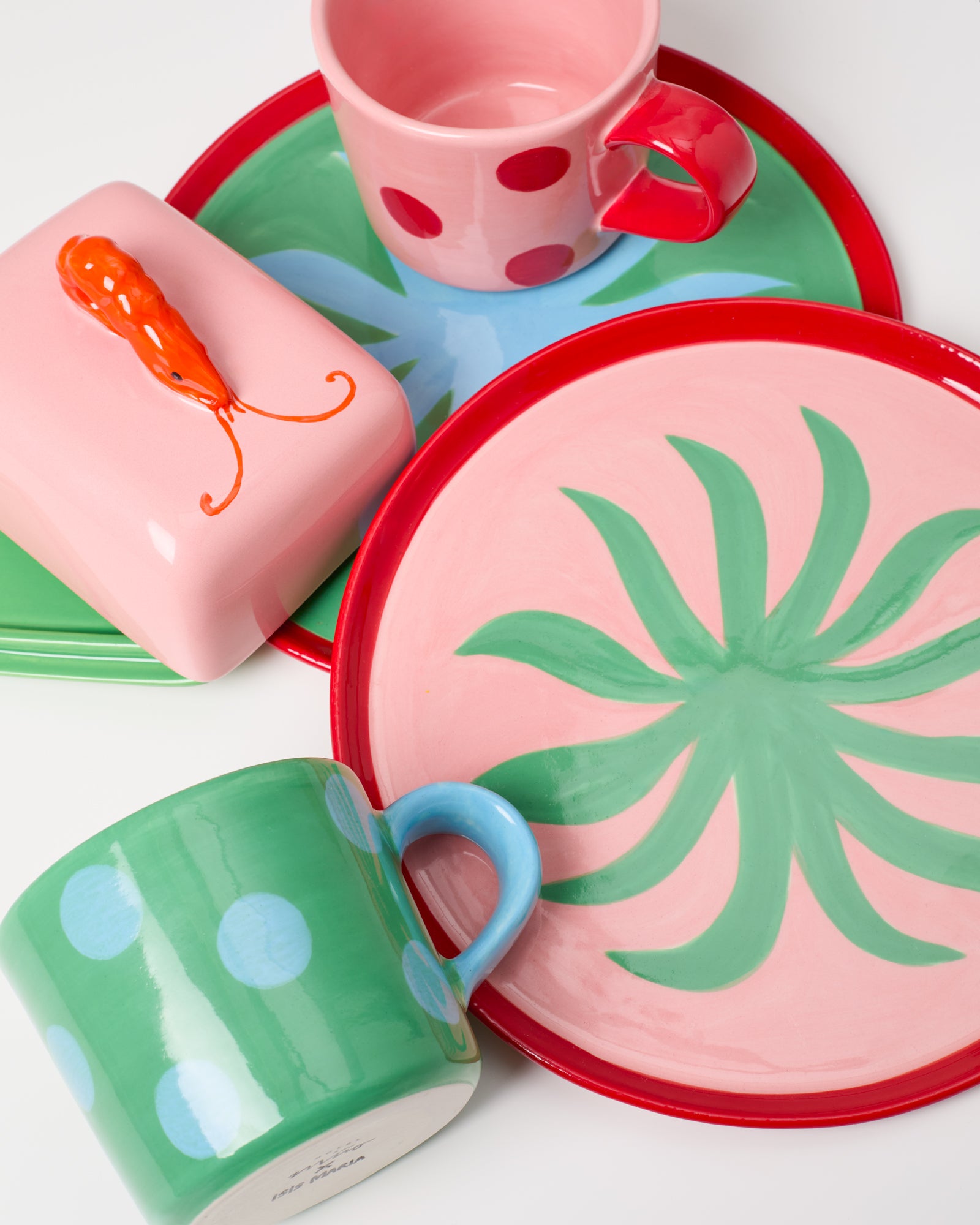 Bunte Keramikteller und Tassen mit roten Punkten, grünen Mustern und eine Schachtel mit orangefarbener Garnelen-Dekoration.