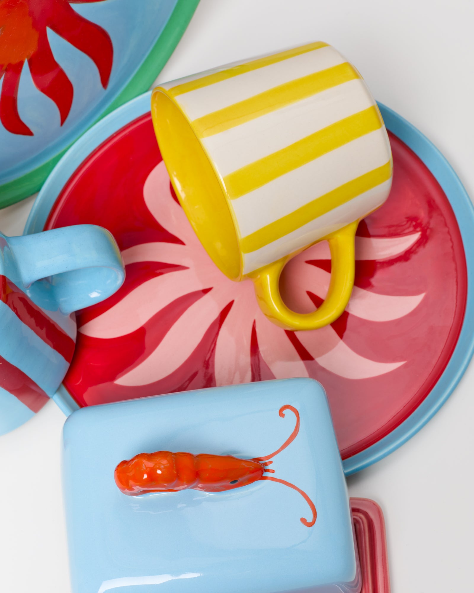 Bunte Keramikgeschirrteile, darunter ein gelber Becher mit Streifen, rote Teller mit Blumenmuster und eine blaue Dose mit Krabbe.