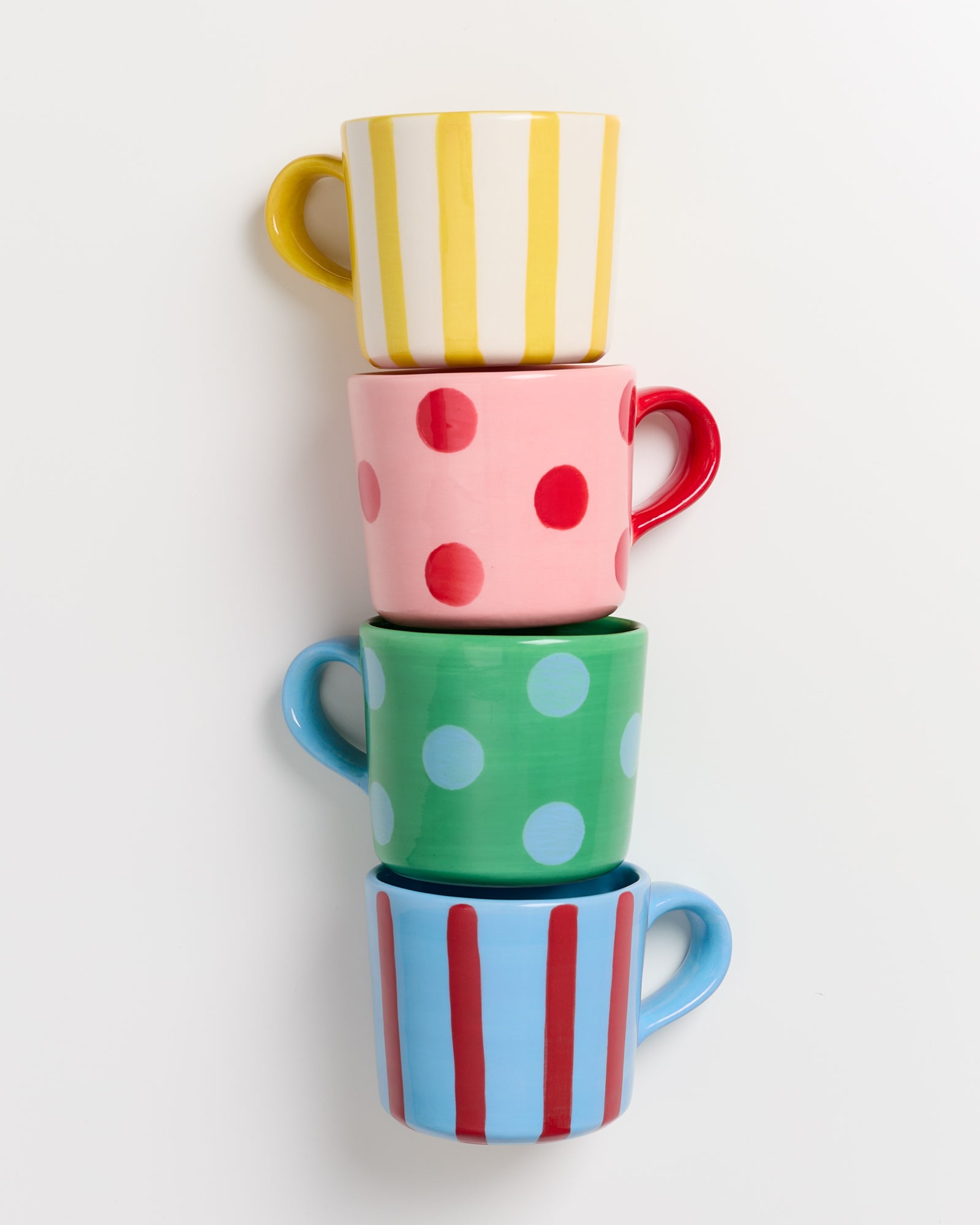 Vier gestapelte Tassen mit bunten Mustern: gelb-weiß gestreift, rosa mit roten Punkten, grün mit blauen Punkten, blau-rot gestreift.