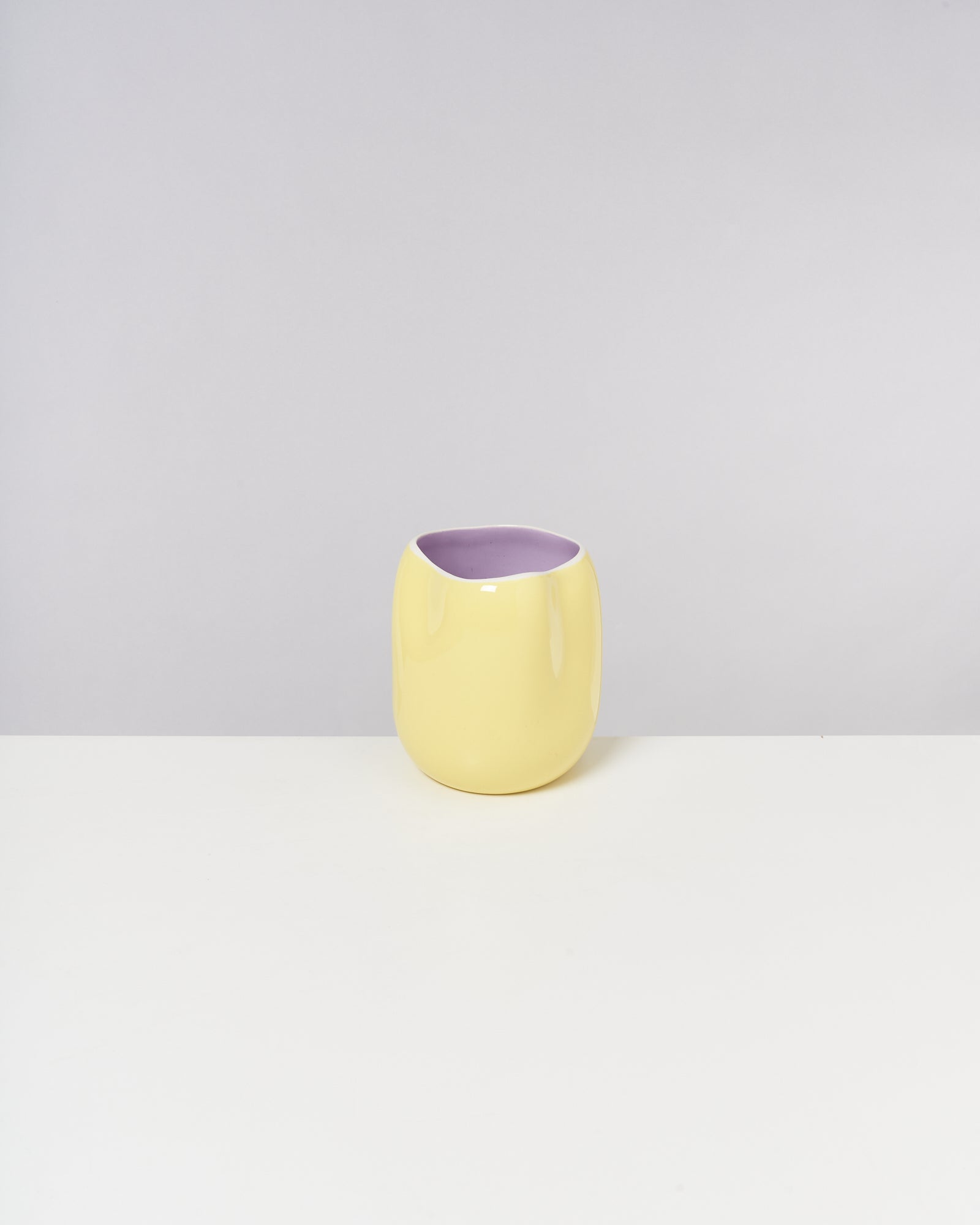 Gelber Keramikbecher mit lila Innenseite auf weißem Tisch vor grauem Hintergrund.