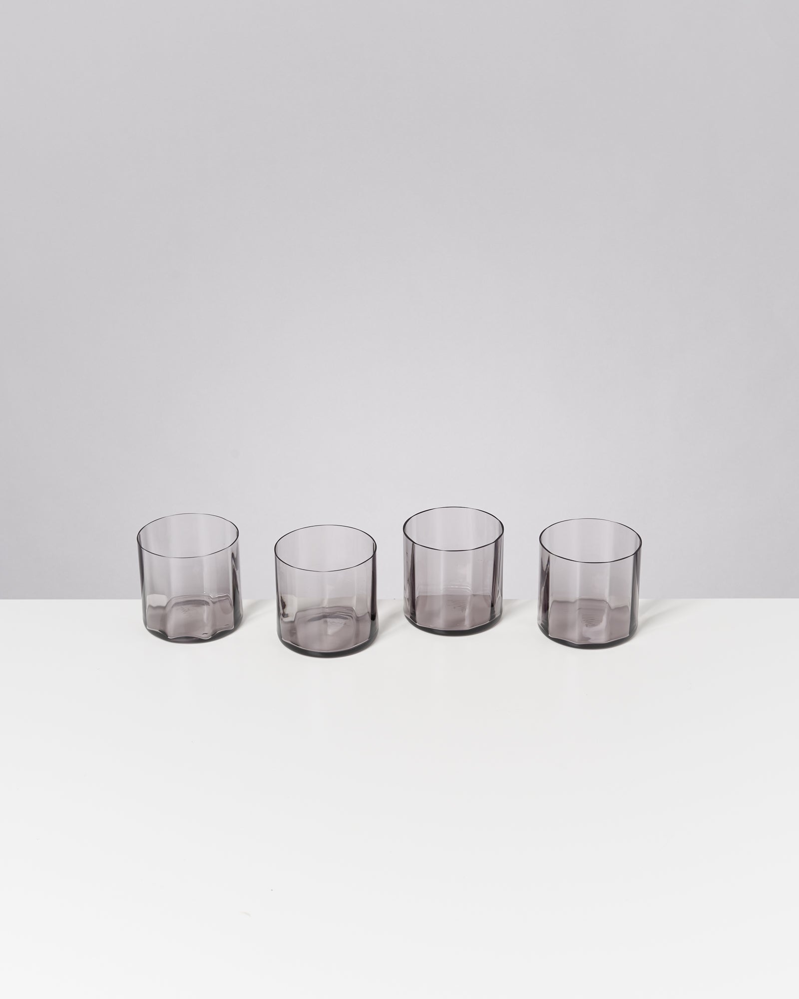 Vier transparente, graue Gläser stehen in einer Reihe auf einem weißen Tisch vor einem grauen Hintergrund.