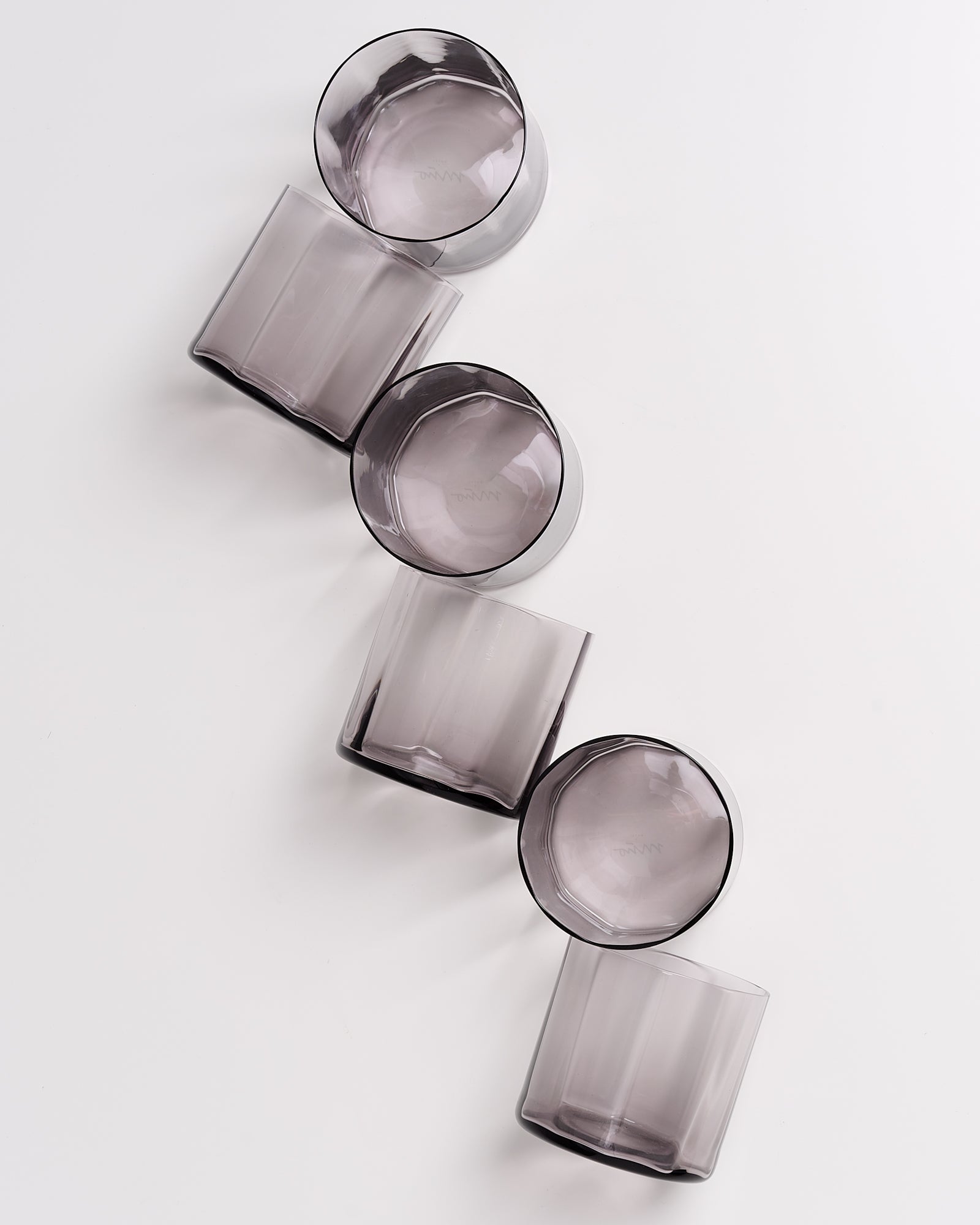 Fünf graue Trinkgläser liegen in einer diagonalen Linie auf einem weißen Hintergrund.