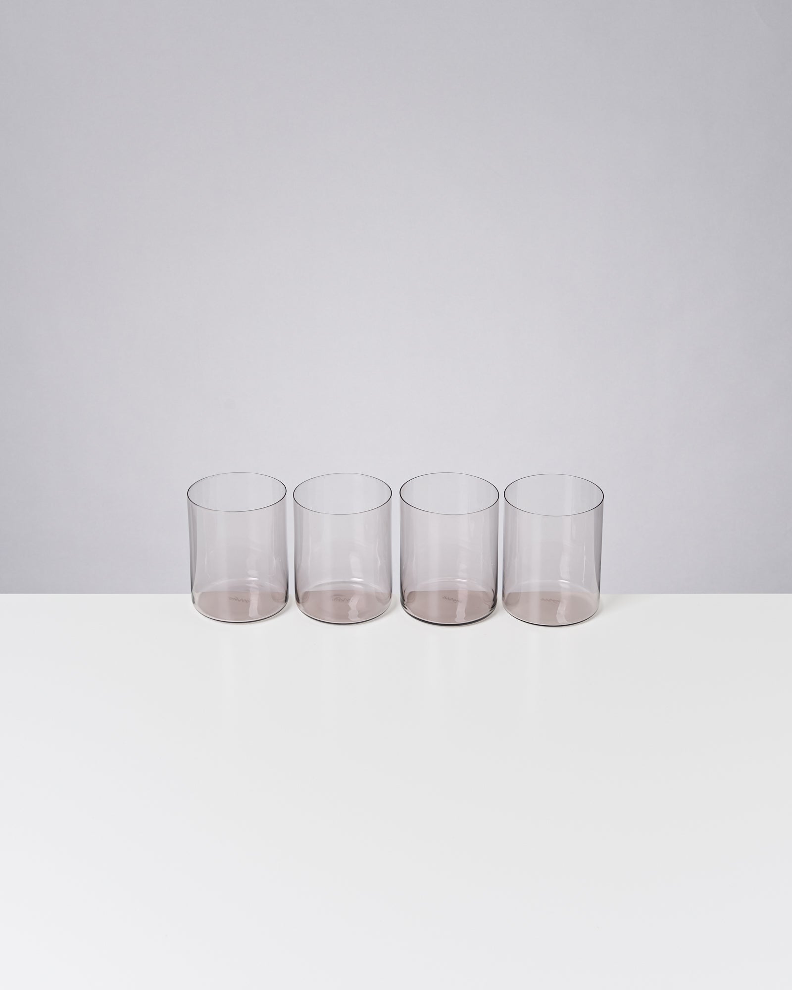 Vier transparente Trinkgläser stehen nebeneinander auf einem weißen Untergrund vor einer grauen Wand.