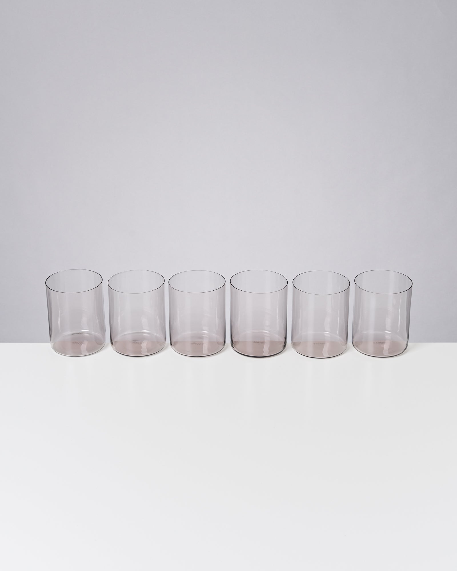 Sechs durchsichtige Trinkgläser stehen in einer Reihe auf einer weißen Oberfläche vor grauem Hintergrund.