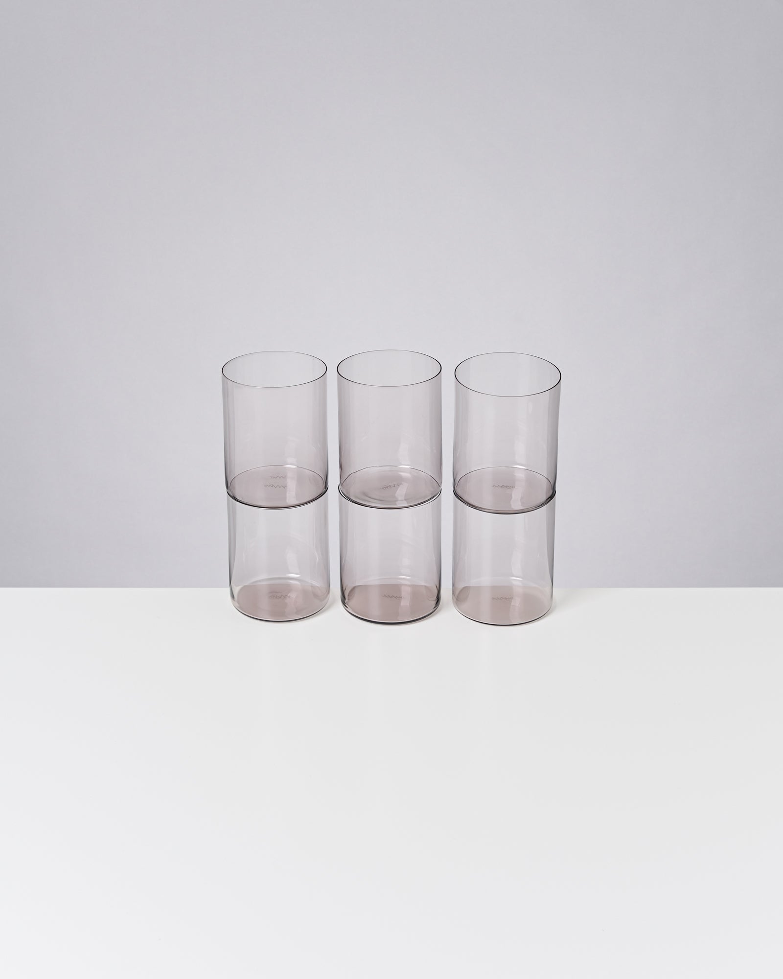 Sechs gestapelte, transparente Becher stehen auf einem weißen Tisch vor grauem Hintergrund.