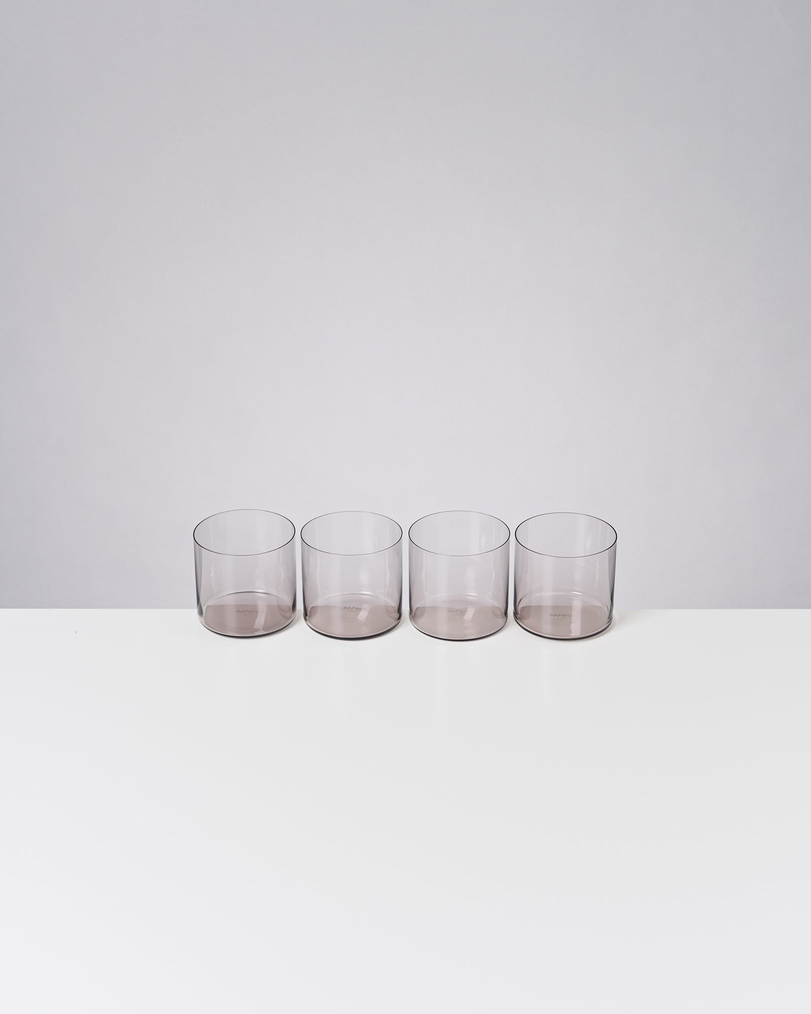 Vier leere, transparente Gläser stehen in einer Reihe auf einem weißen Tisch vor einem grauen Hintergrund.