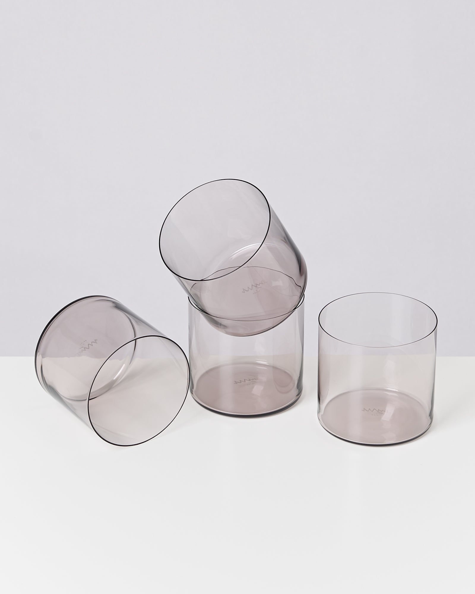 Vier durchsichtige, graue Gläser liegen und stehen auf einem weißen Tisch vor einem hellgrauen Hintergrund.