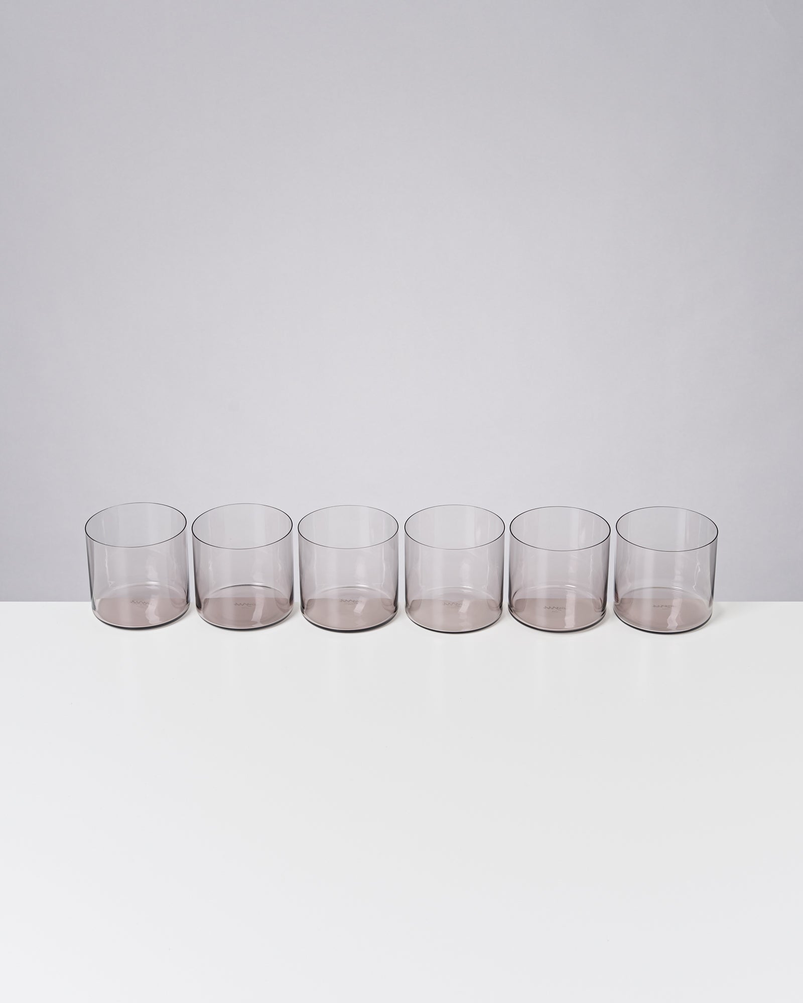 Sechs leere, transparente Gläser stehen in einer Reihe auf einem weißen Tisch.