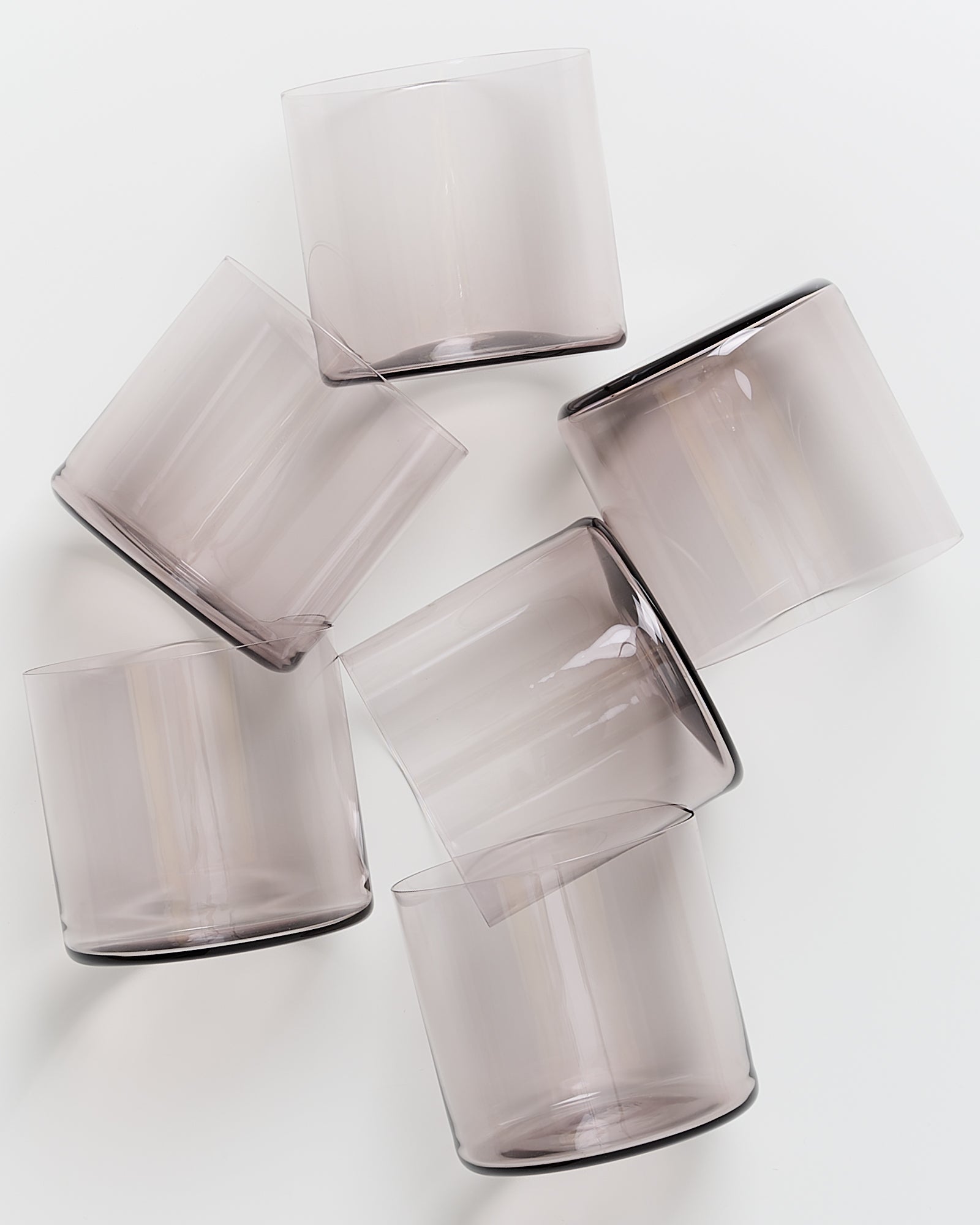 Sechs transparente, leicht getönte Trinkgläser liegen schräg übereinander auf einer weißen Oberfläche.