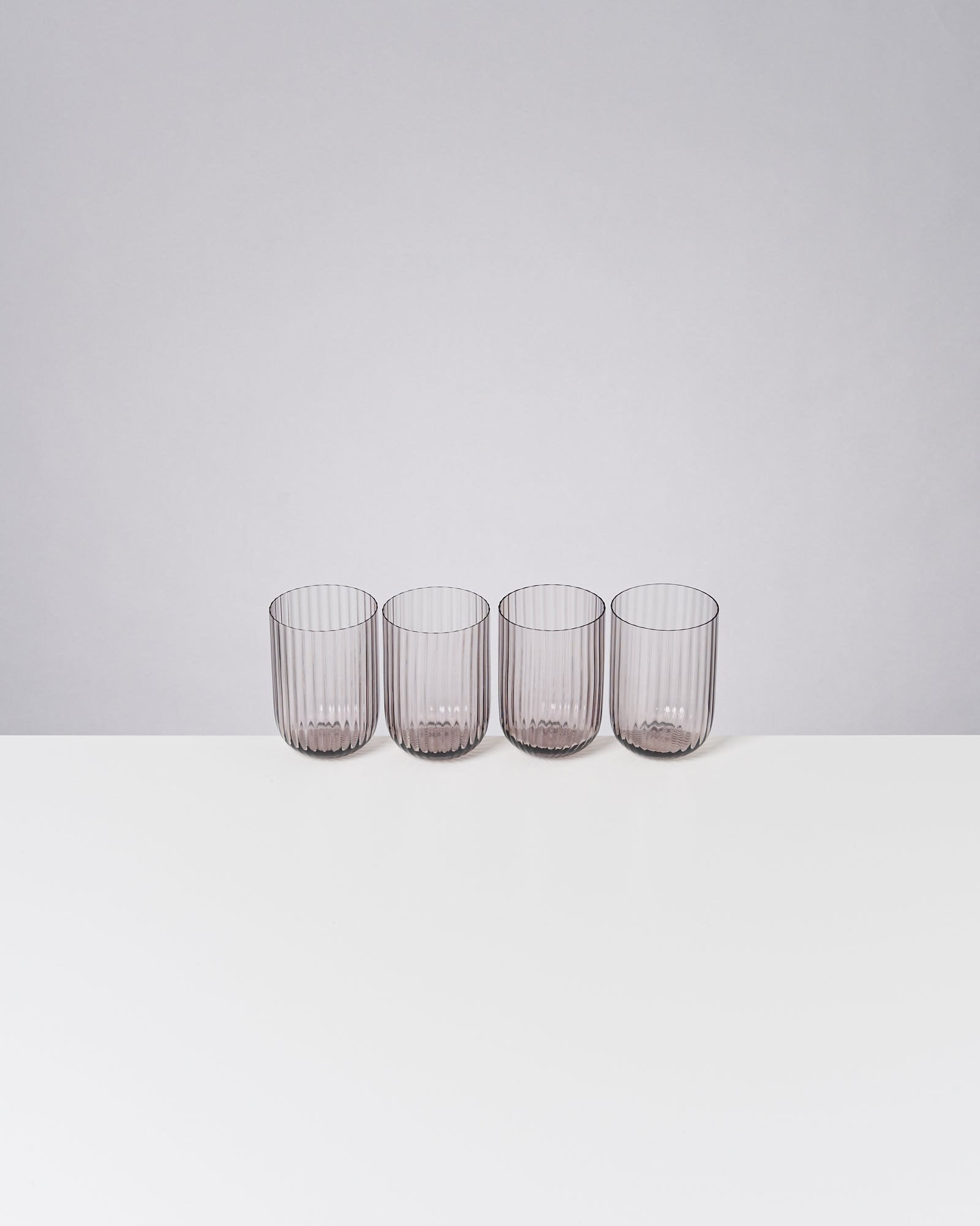 Vier gerippte Gläser stehen nebeneinander auf einer weißen Oberfläche vor einem hellgrauen Hintergrund.