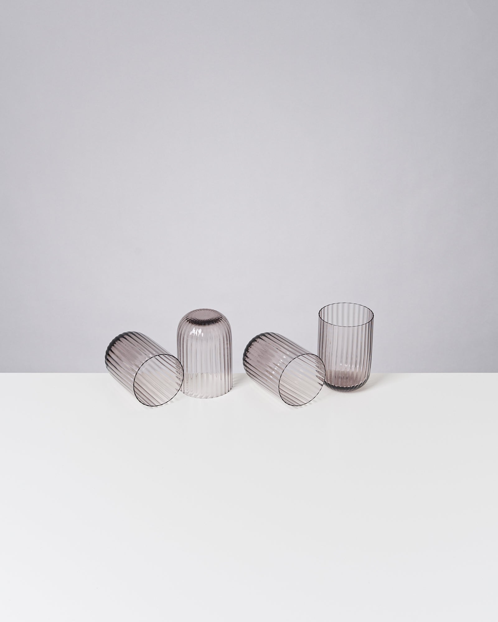 Vier geriffelte Gläser, drei liegen und eines steht, auf einem weißen Tisch vor grauem Hintergrund.