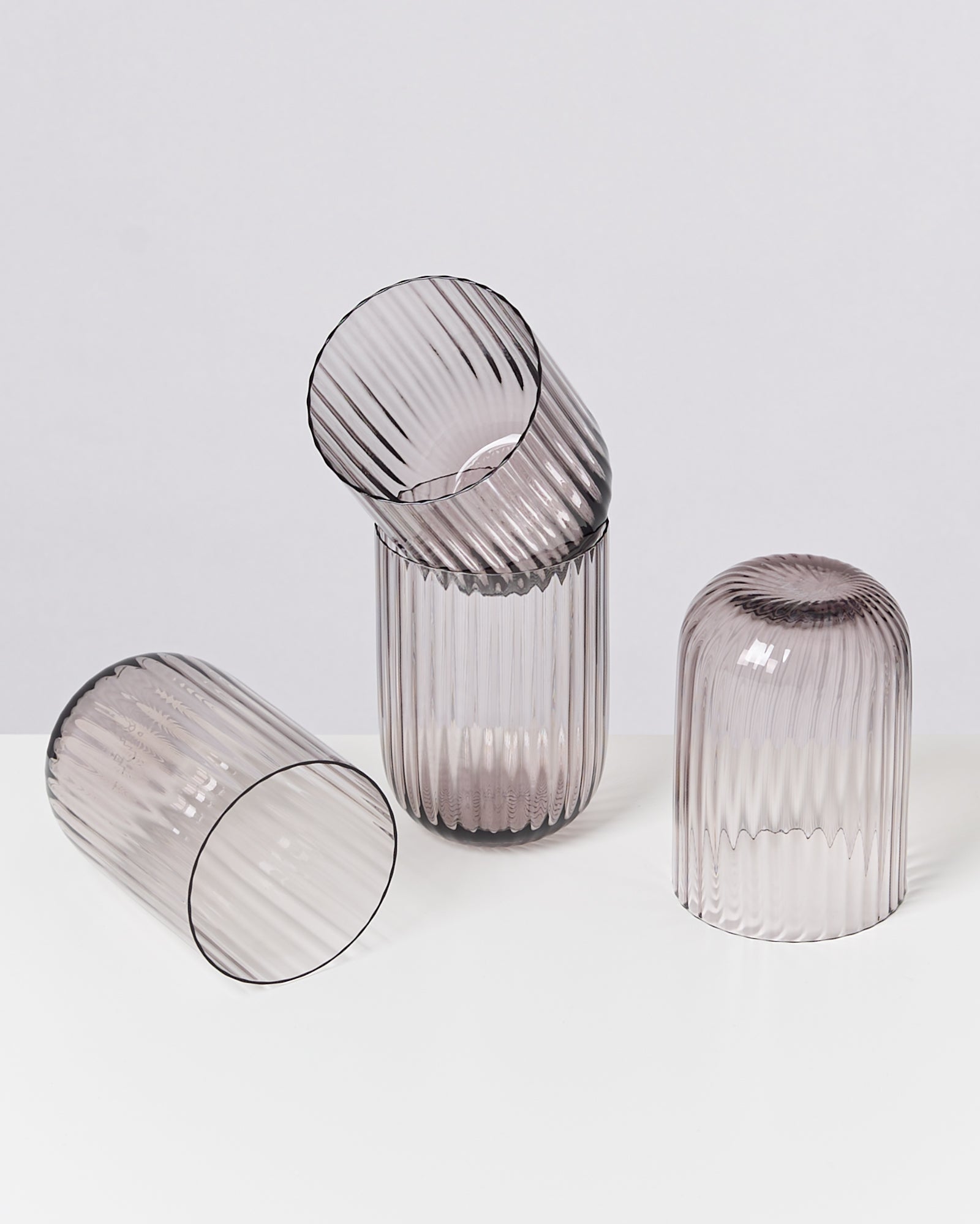 Drei geriffelte, rauchgraue Glasvasen auf einem weißen Tisch, eine liegt, zwei stehen senkrecht und kopfüber.