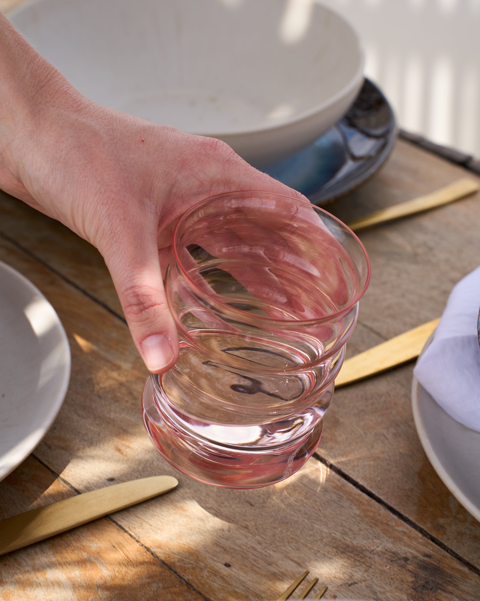Eine Hand hält ein rosa Glas über einem gedeckten Holztisch mit goldenem Besteck und Keramiktellern.