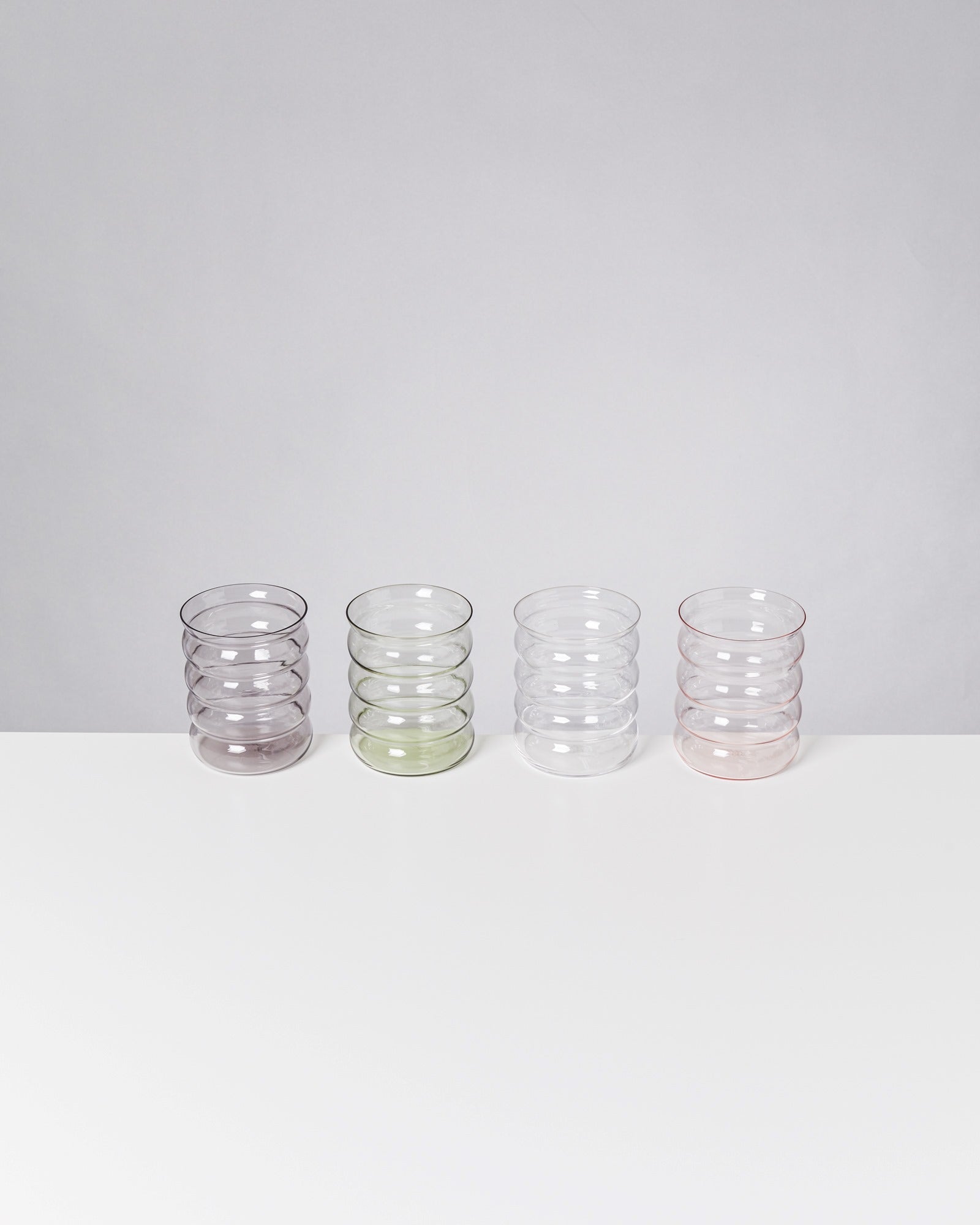 Vier gestapelte, durchsichtige Gläser in unterschiedlichen Farben stehen nebeneinander auf einem weißen Tisch.