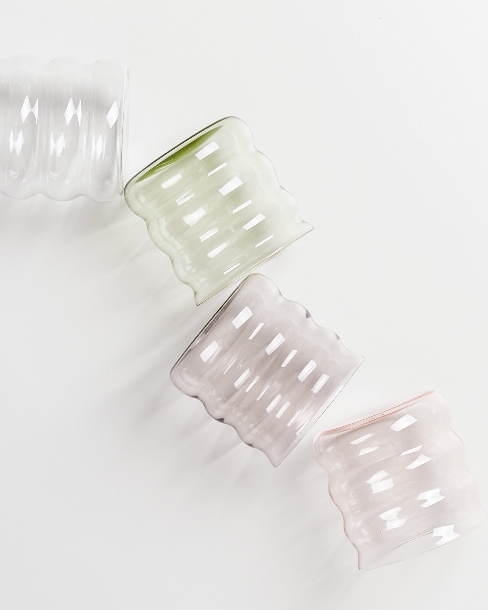 Vier gewellte, transparente Gläser in verschiedenen Pastellfarben liegen diagonal auf einer weißen Oberfläche.
