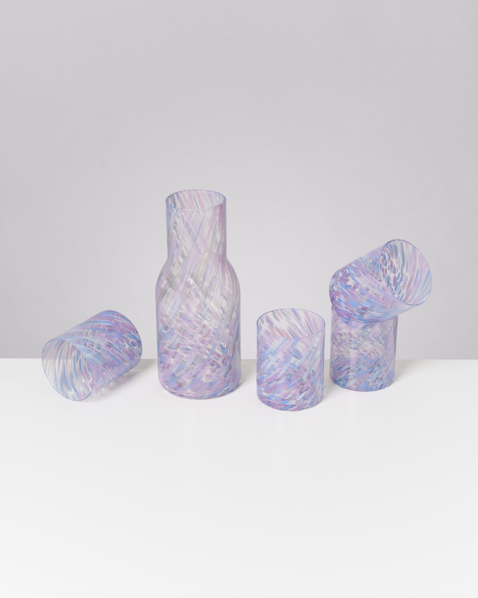 Vier bunte Gläser und eine Karaffe aus gemustertem Glas stehen auf einem weißen Tisch vor einem grauen Hintergrund.