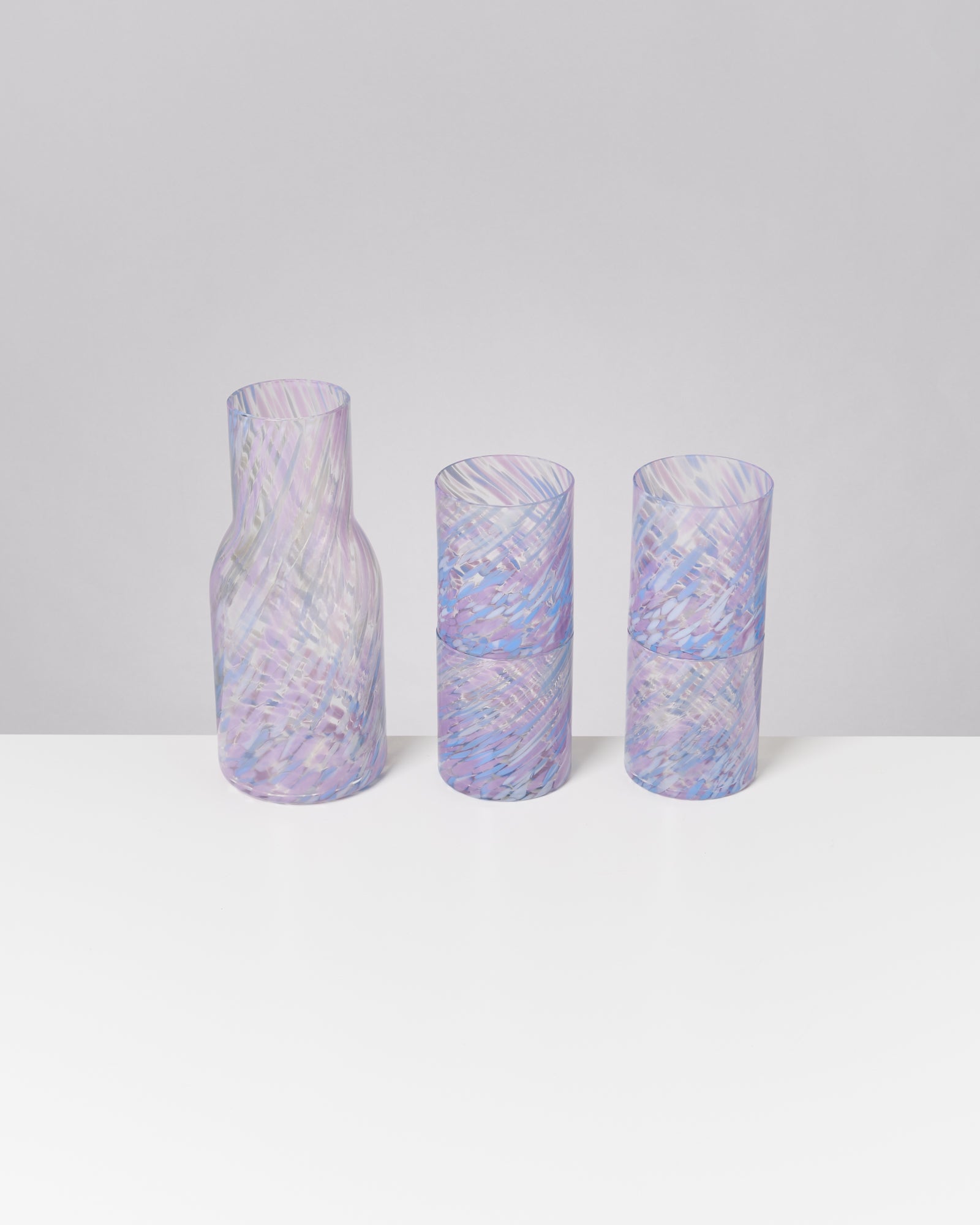 Violette und blaue Glaskaraffe mit zwei passenden Gläsern auf einem weißen Tisch vor einem grauen Hintergrund.