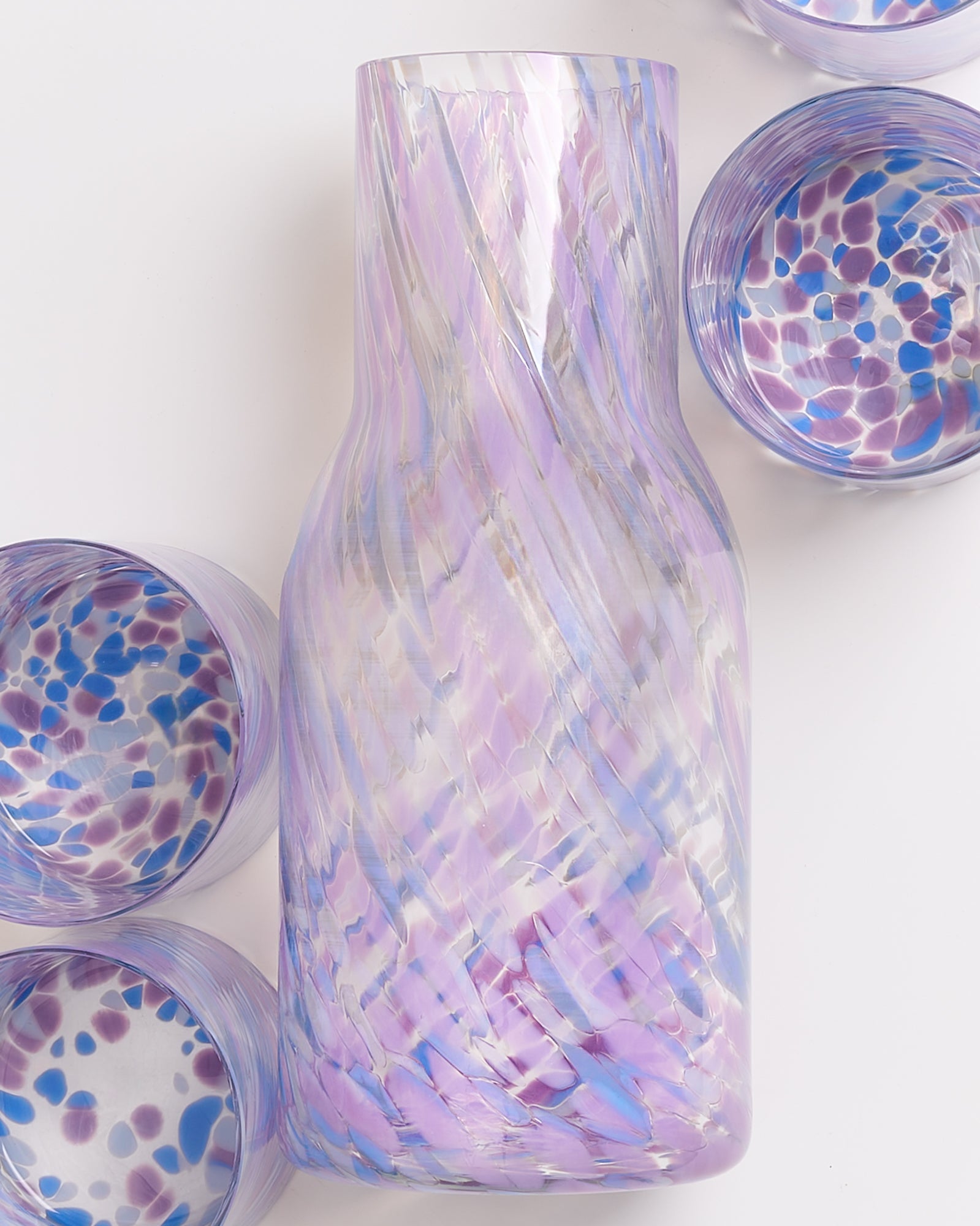 Eine glasierte Glaskaraffe mit lila Wirbeln, umgeben von passenden Gläsern mit blauen und lila Punkten.