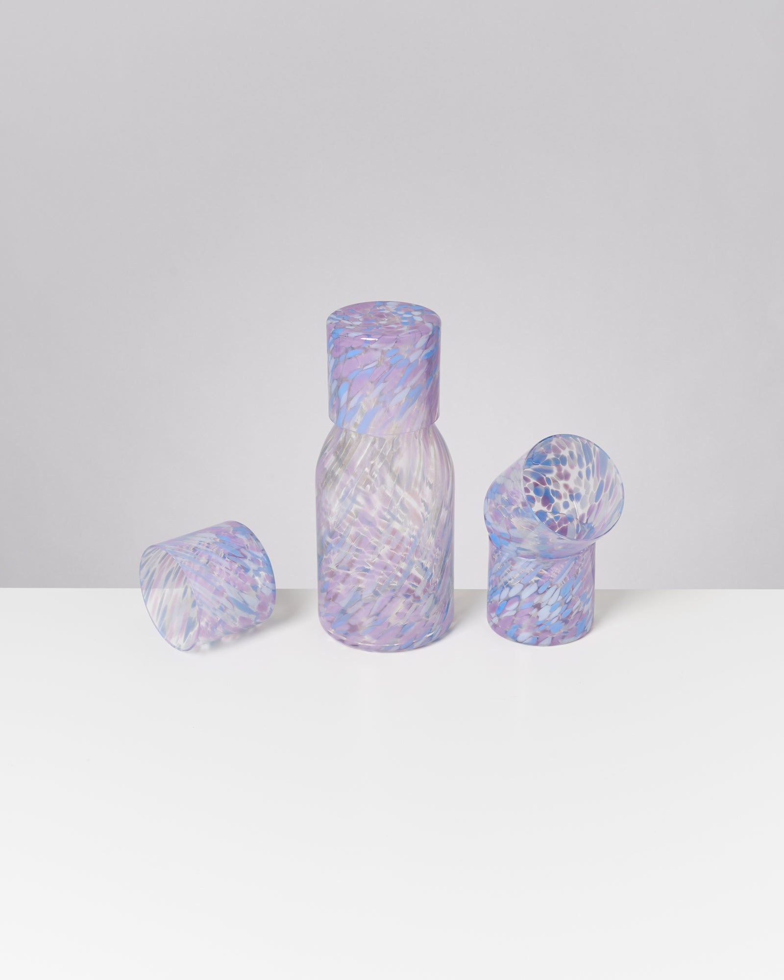 Drei bunte Glasgefäße in Lila- und Blautönen stehen auf einer weißen Oberfläche vor grauem Hintergrund.